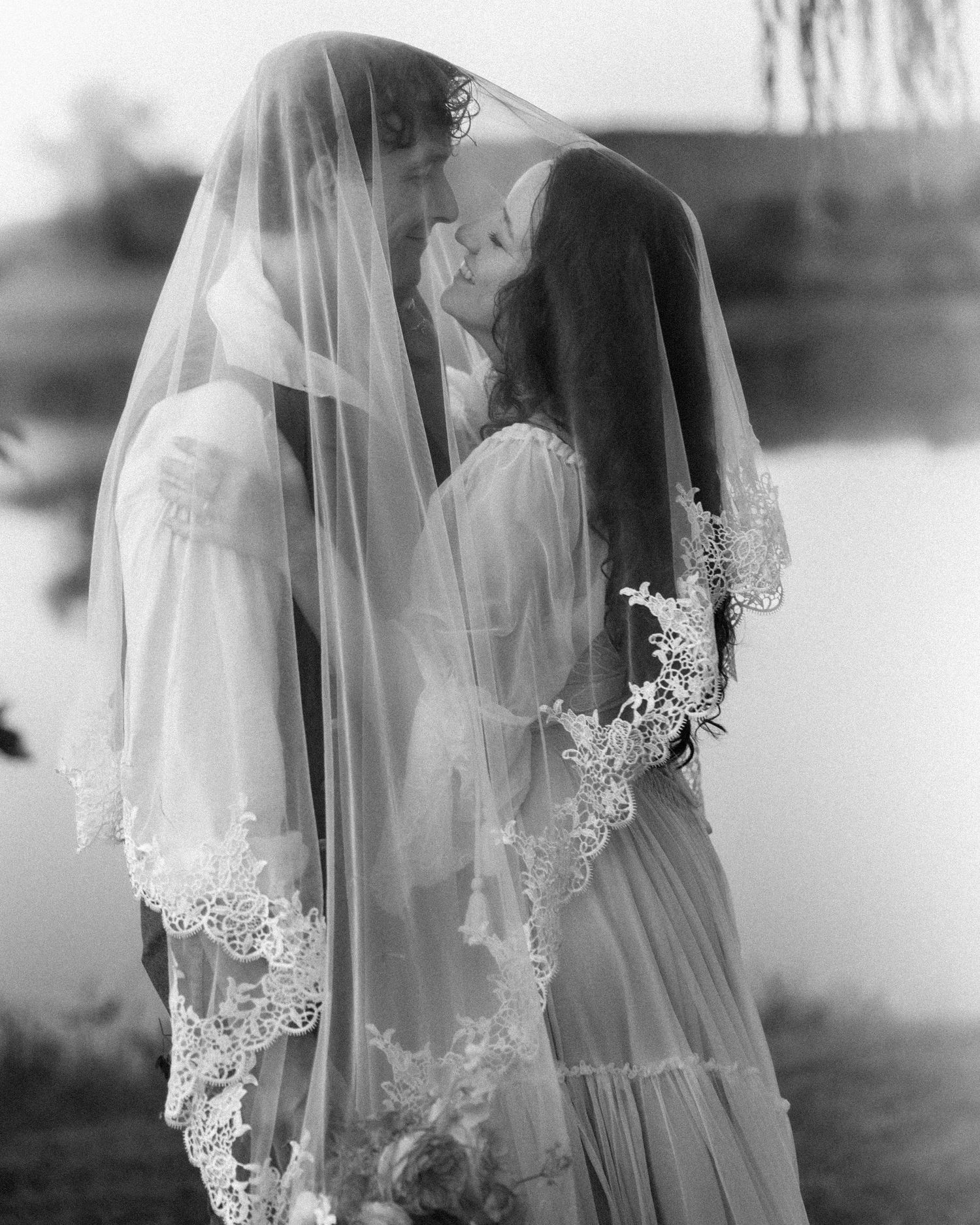 A romantic black and white photograph of a couple in wedding attire sharing an intimate moment under a delicate lace veil.