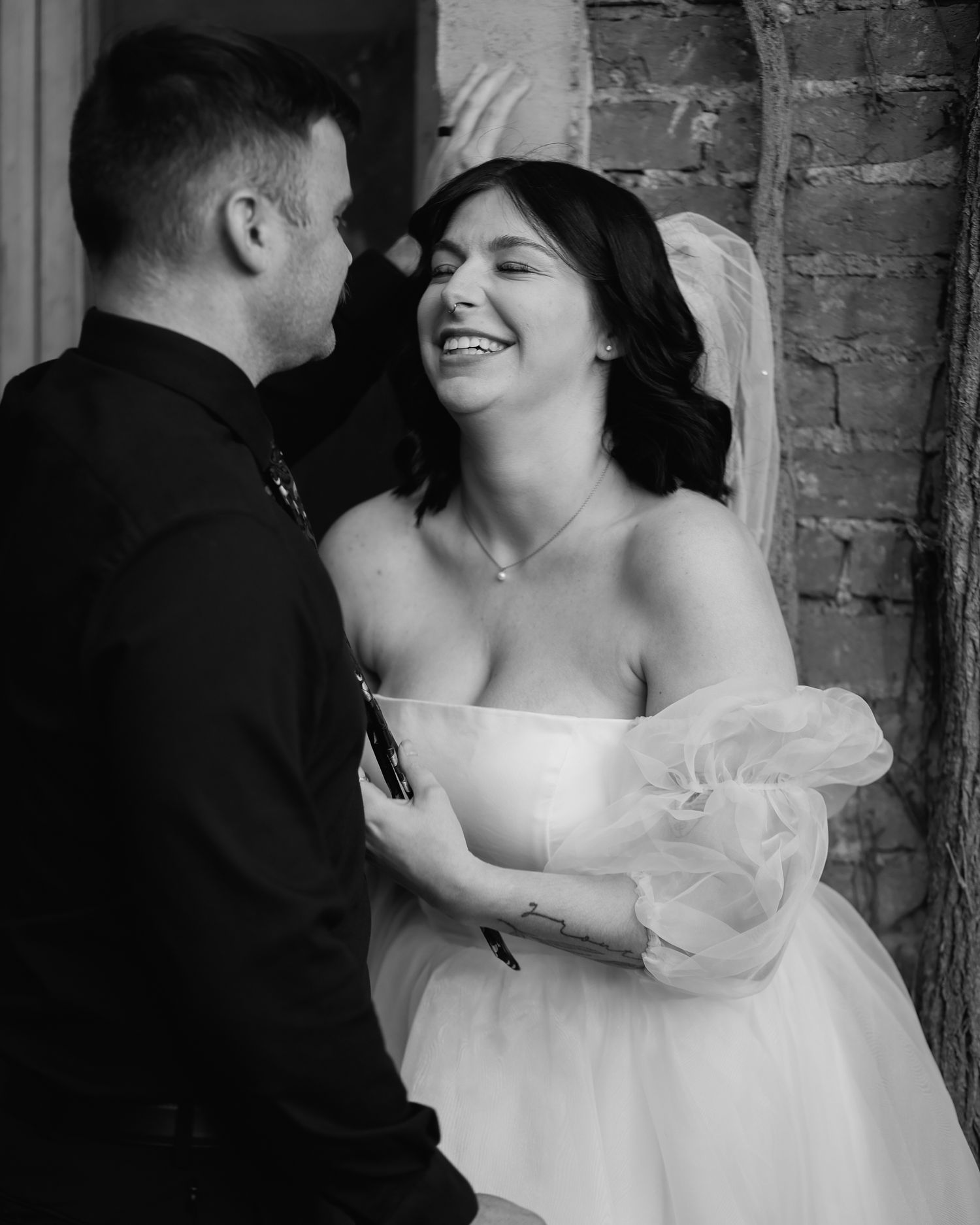 A black and white wedding portrait of a bride and groom sharing an intimate moment against a brick wall.