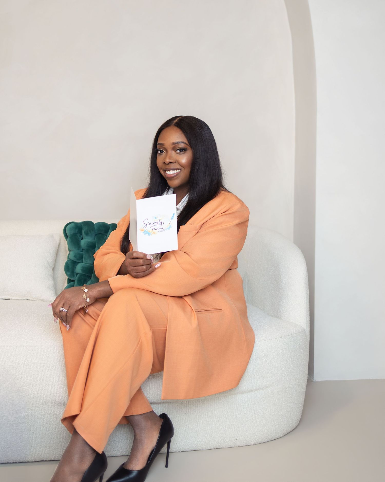 A woman in an orange pantsuit sits on a white sofa, smiling and holding a white card in a bright studio.