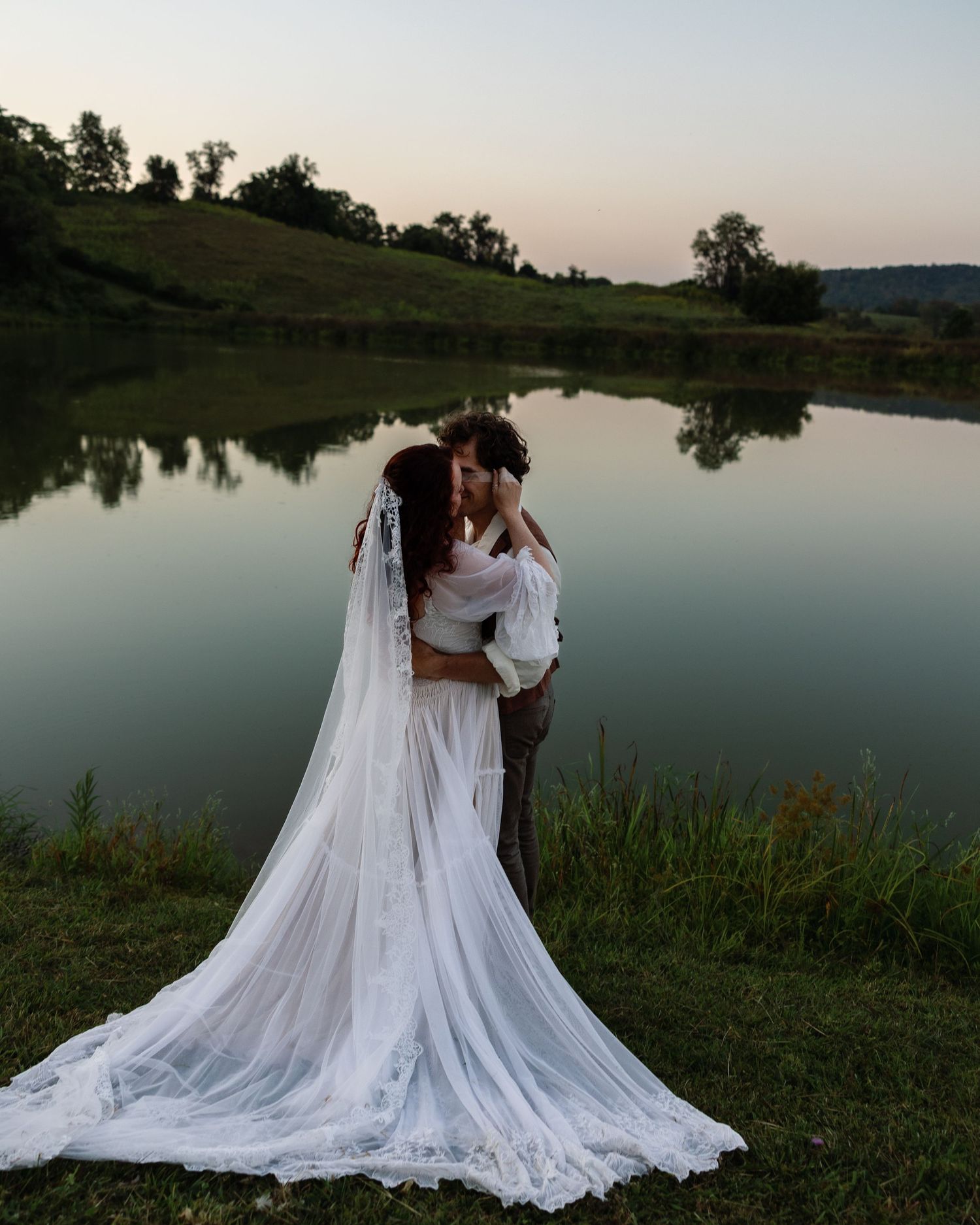 A romantic wedding couple embraces by a serene lake at sunset with a flowing white veil trailing behind them.