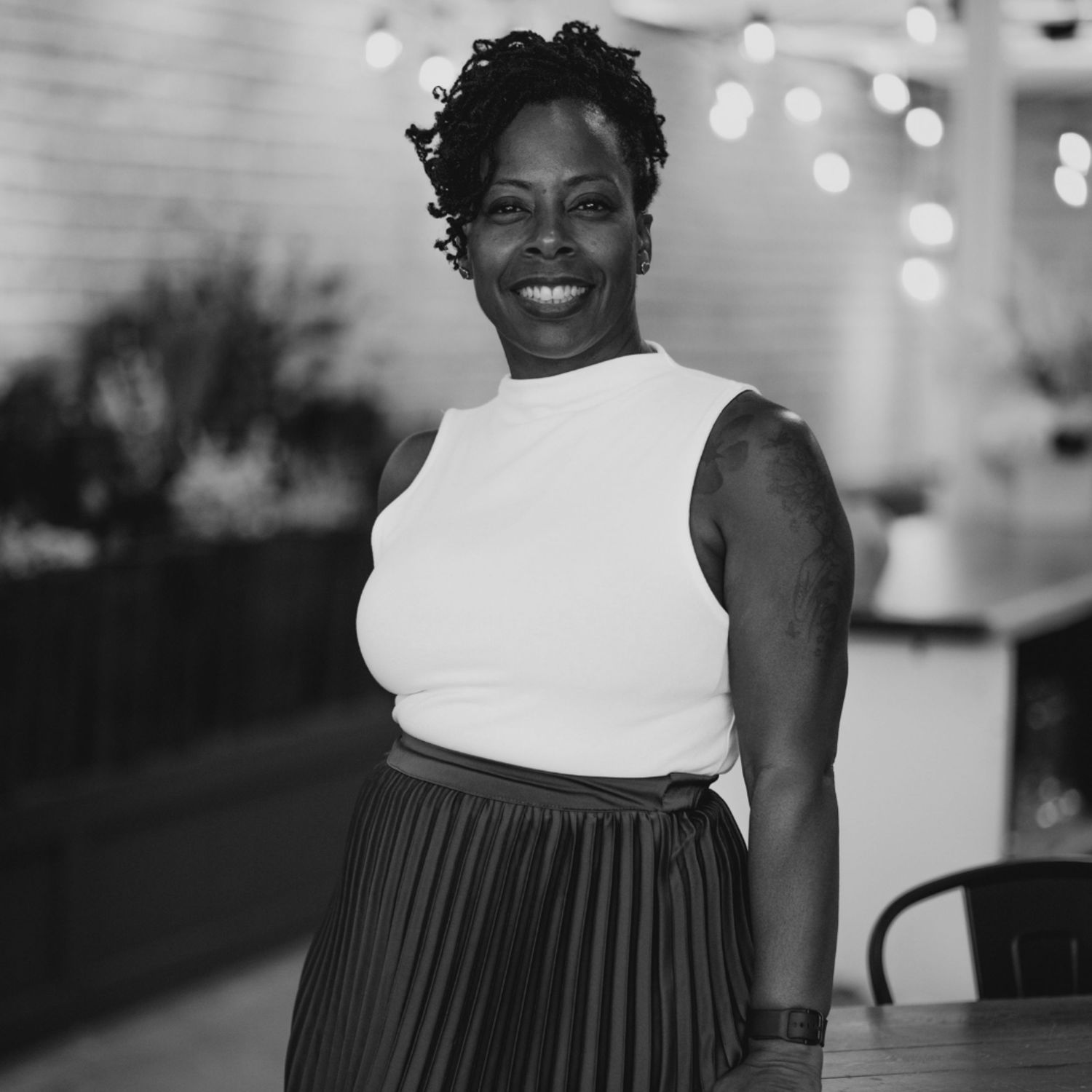 A person in a white sleeveless top and striped skirt smiles warmly against a blurred background with bokeh lights.