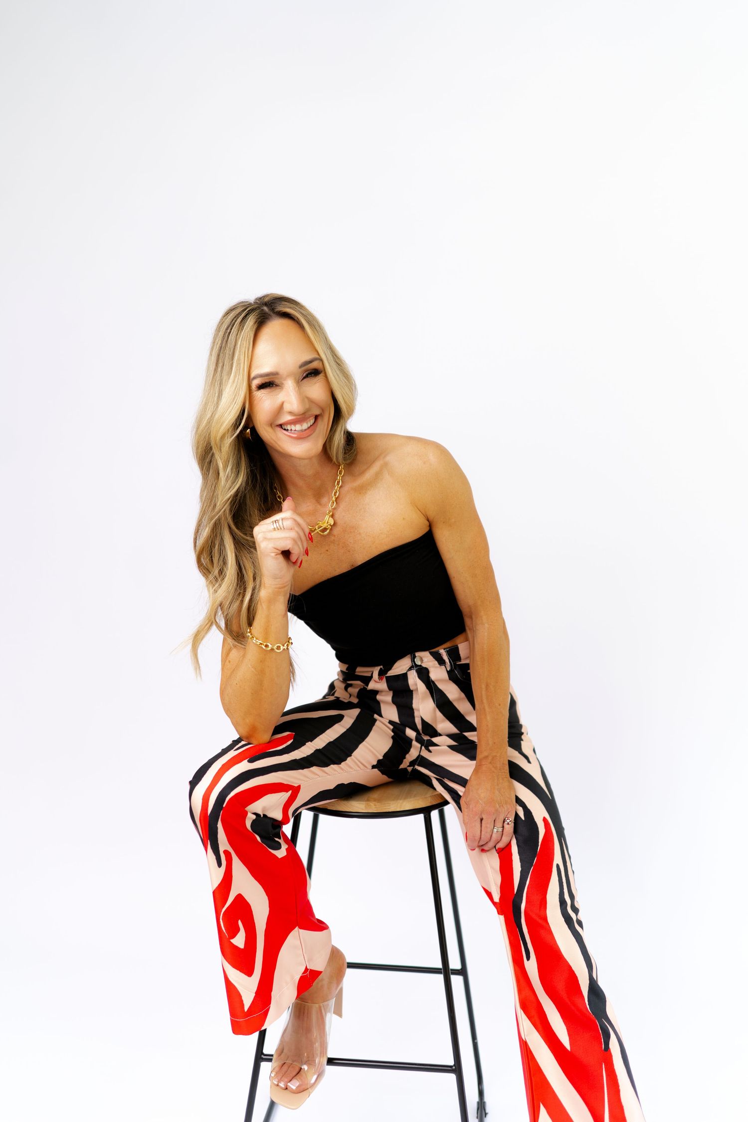Smiling blonde woman seated on stool in black tube top and red abstract print wide-leg pants with gold jewelry.
