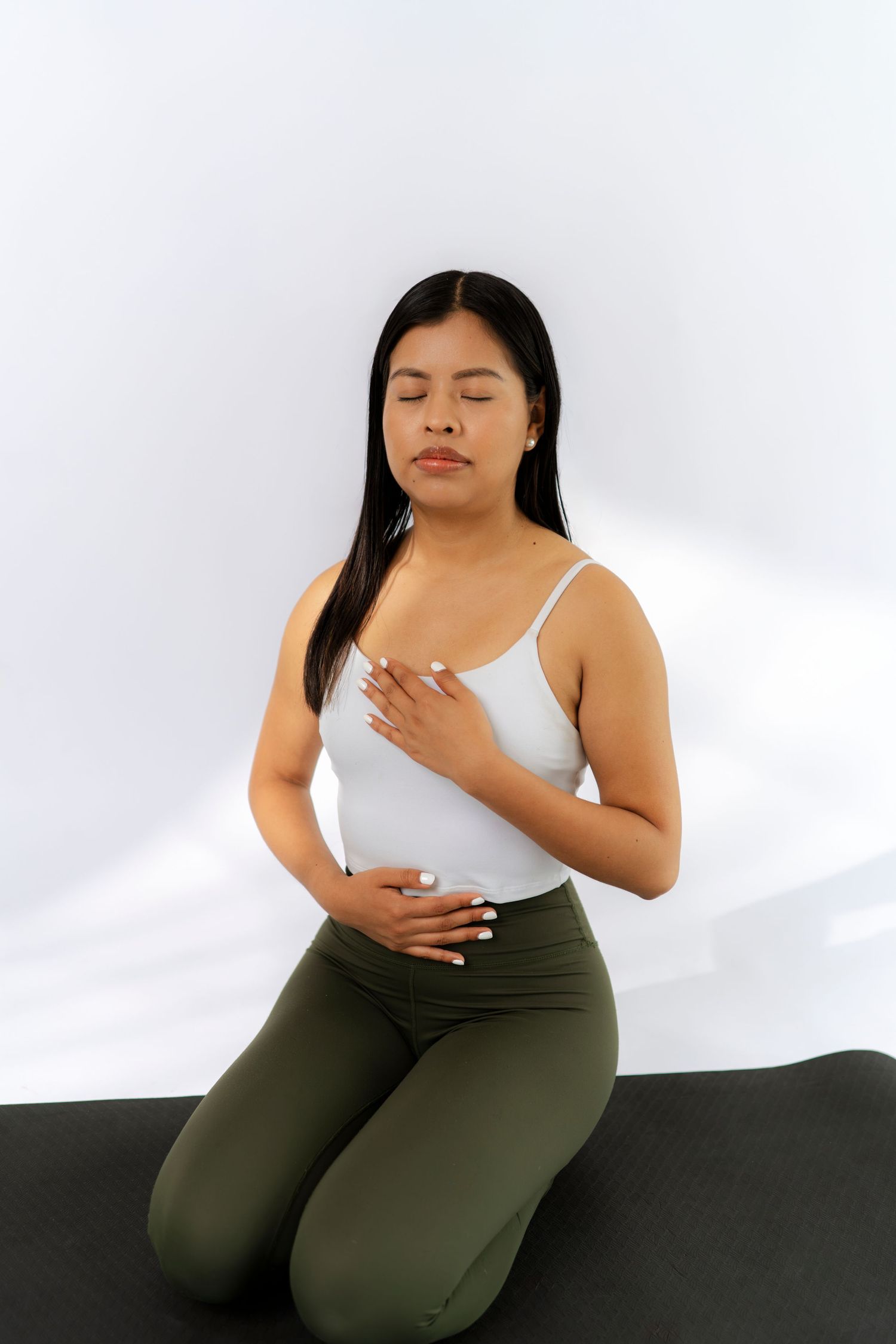 Woman in white tank top and green leggings kneeling on yoga mat with eyes closed, hands on chest.