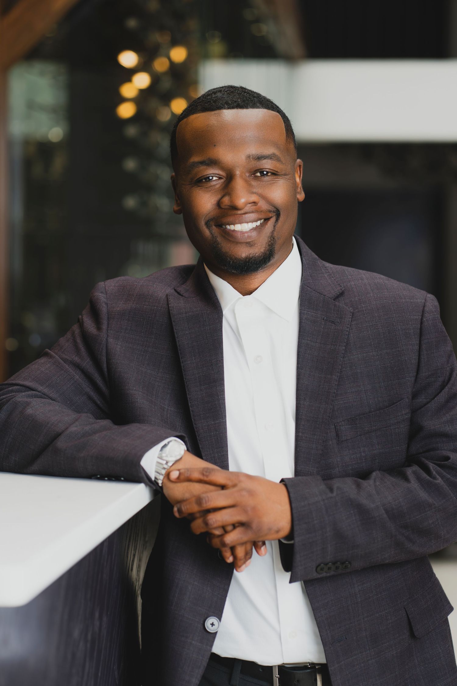 Smiling Black businessman in gray blazer and white shirt, leaning on counter with hands clasped, wearing a silver watch.