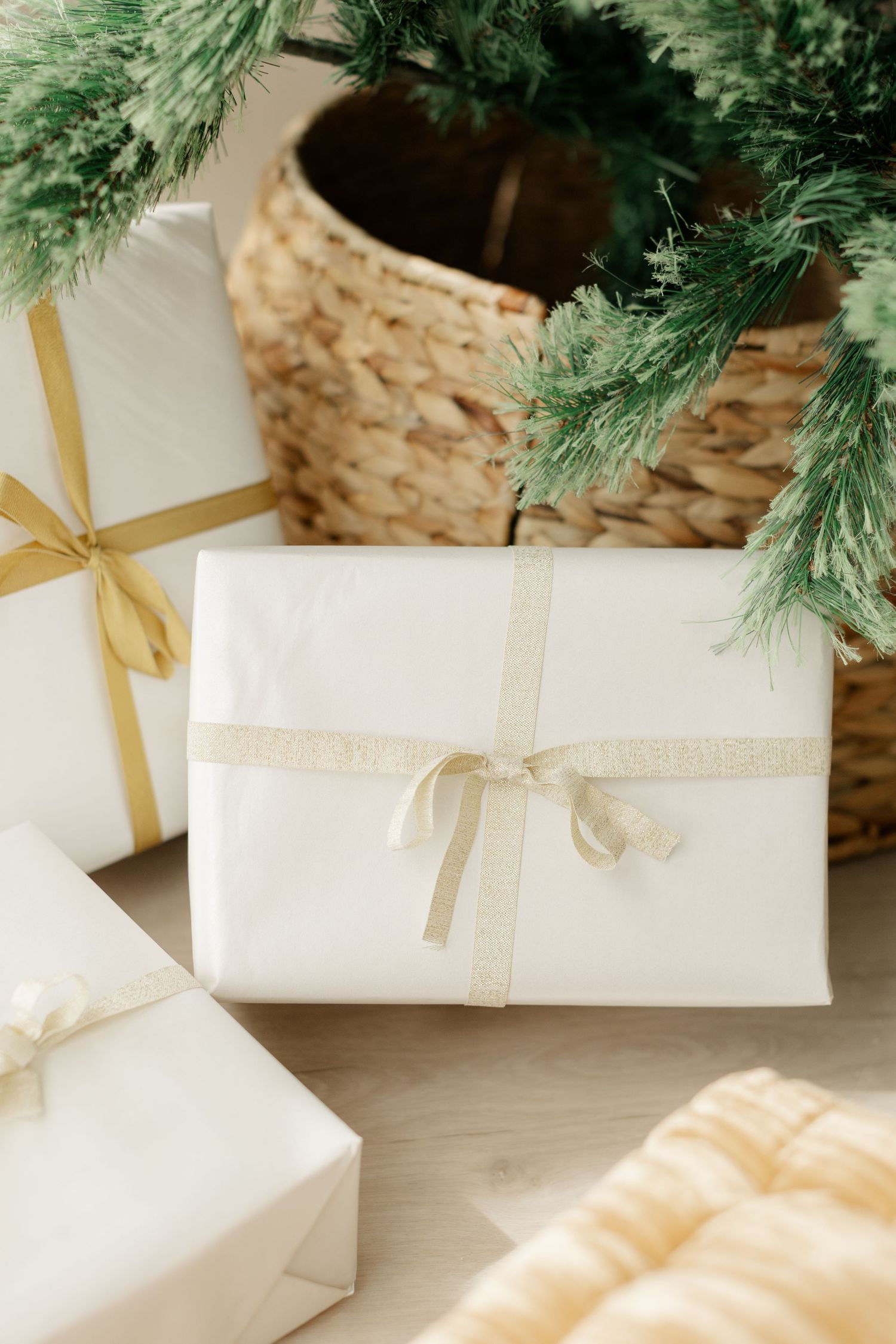 White gift boxes with gold ribbons arranged under a Christmas tree in a wicker basket on a light wood floor.