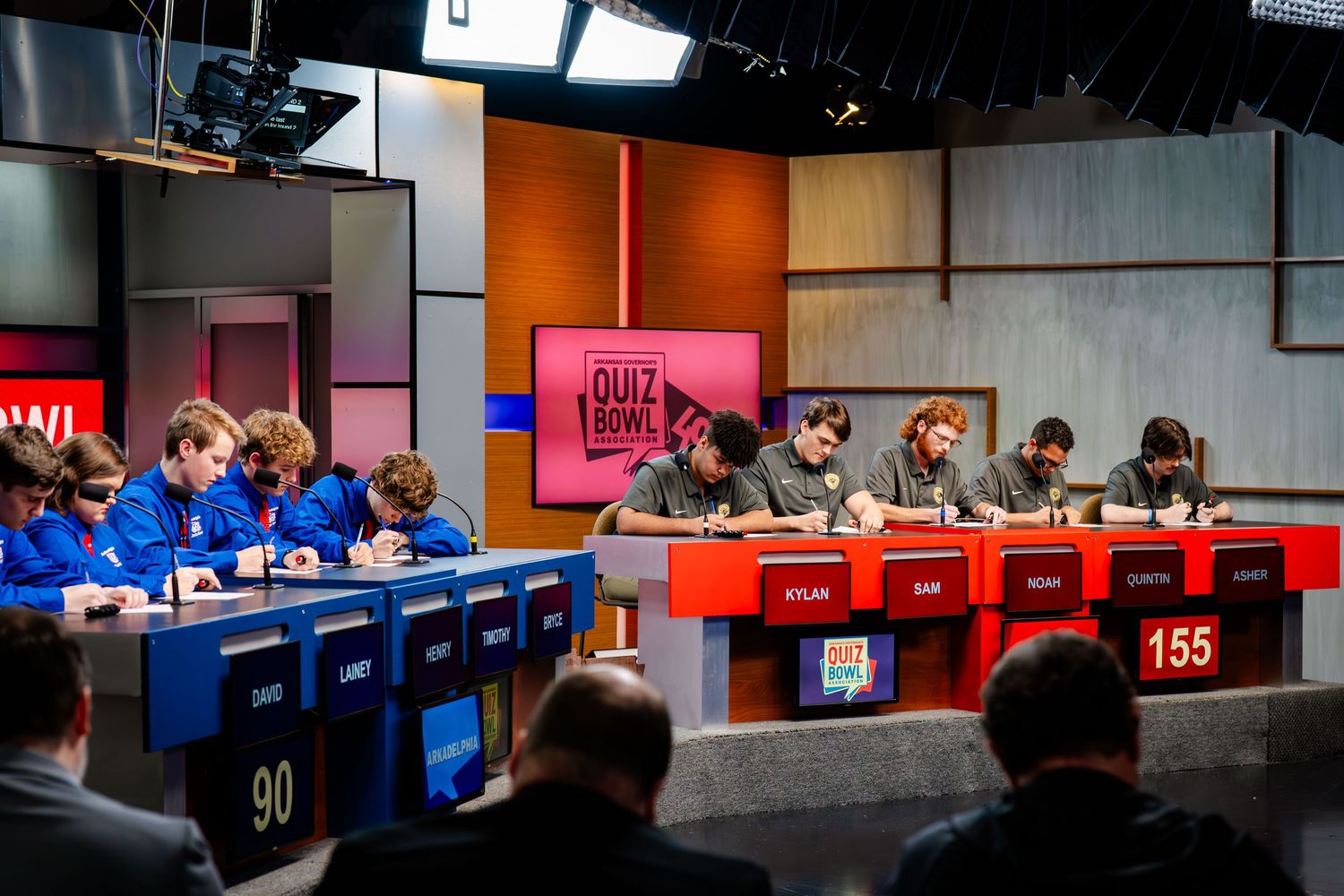 Wide shot of a game show set with blue podiums and orange wall accents.