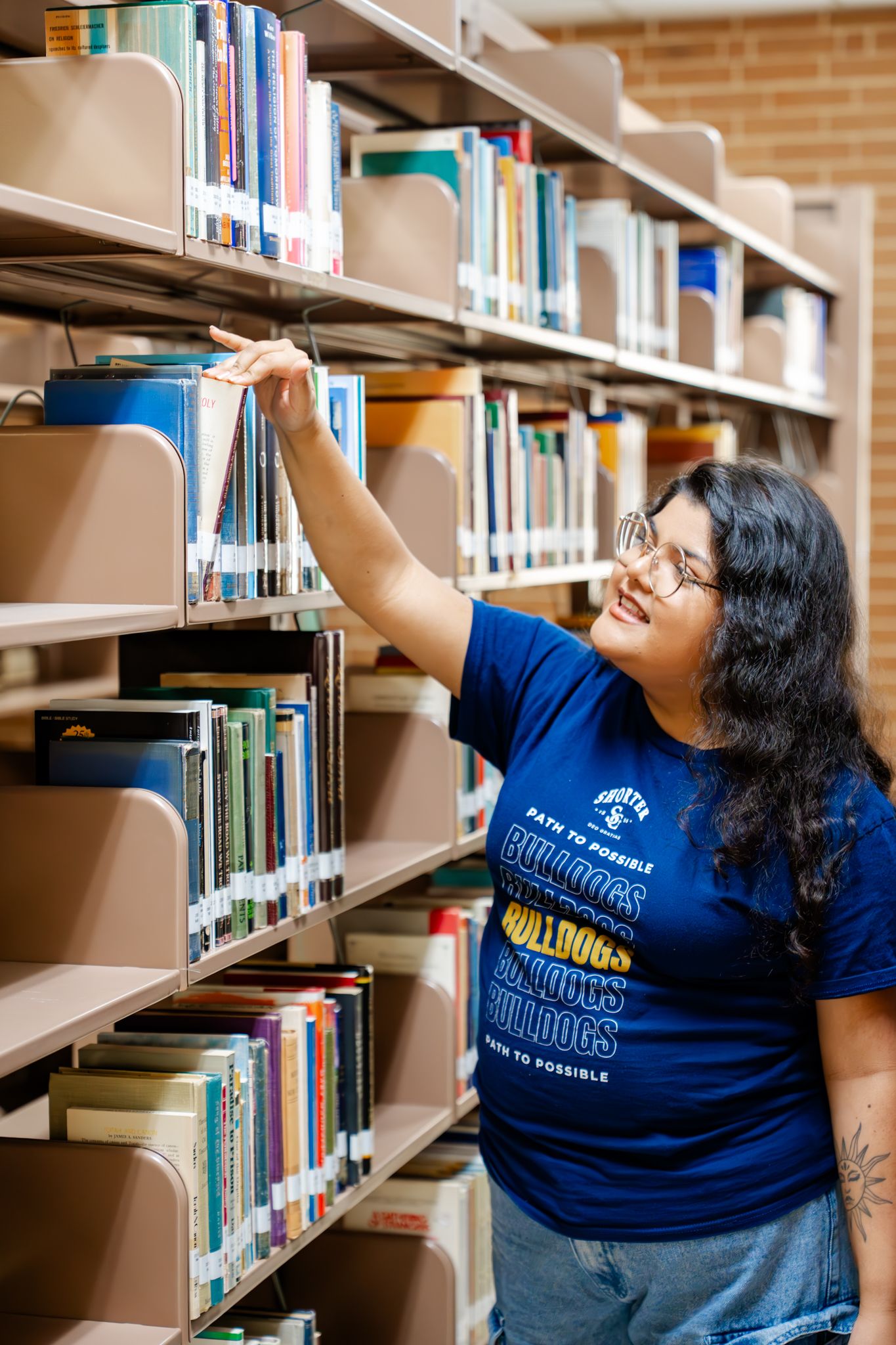 Student browsing books on library shelves while wearing university apparel.