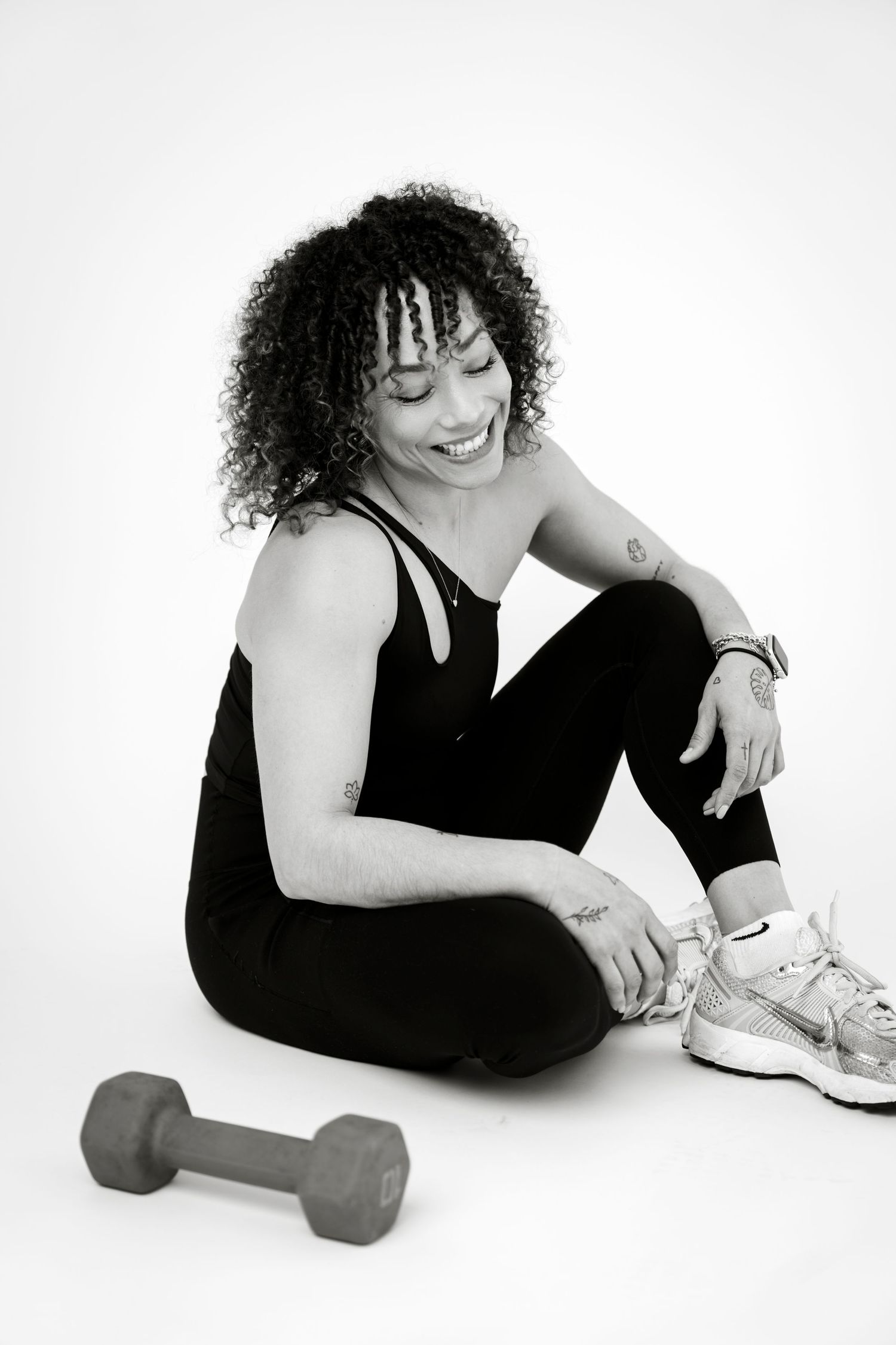 Smiling woman in black athletic wear sitting on floor next to a dumbbell in black and white photo.
