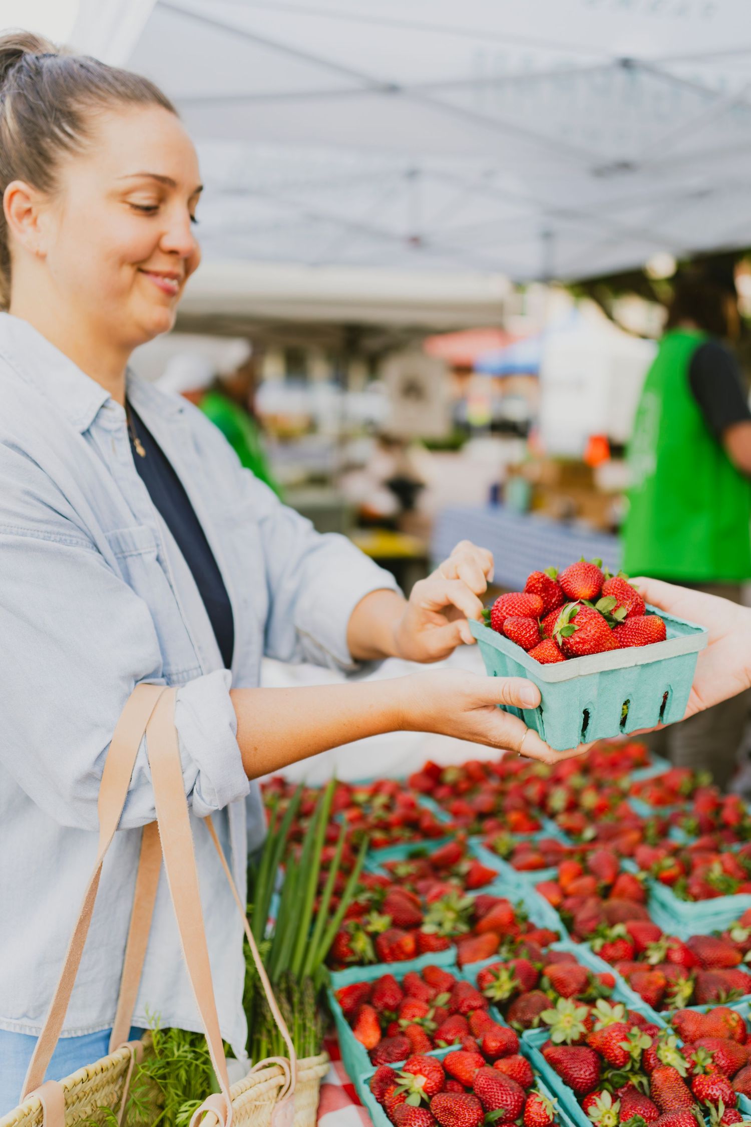 Woman smiling while selecting fresh strawberries in green cartons at an outdoor farmers market.