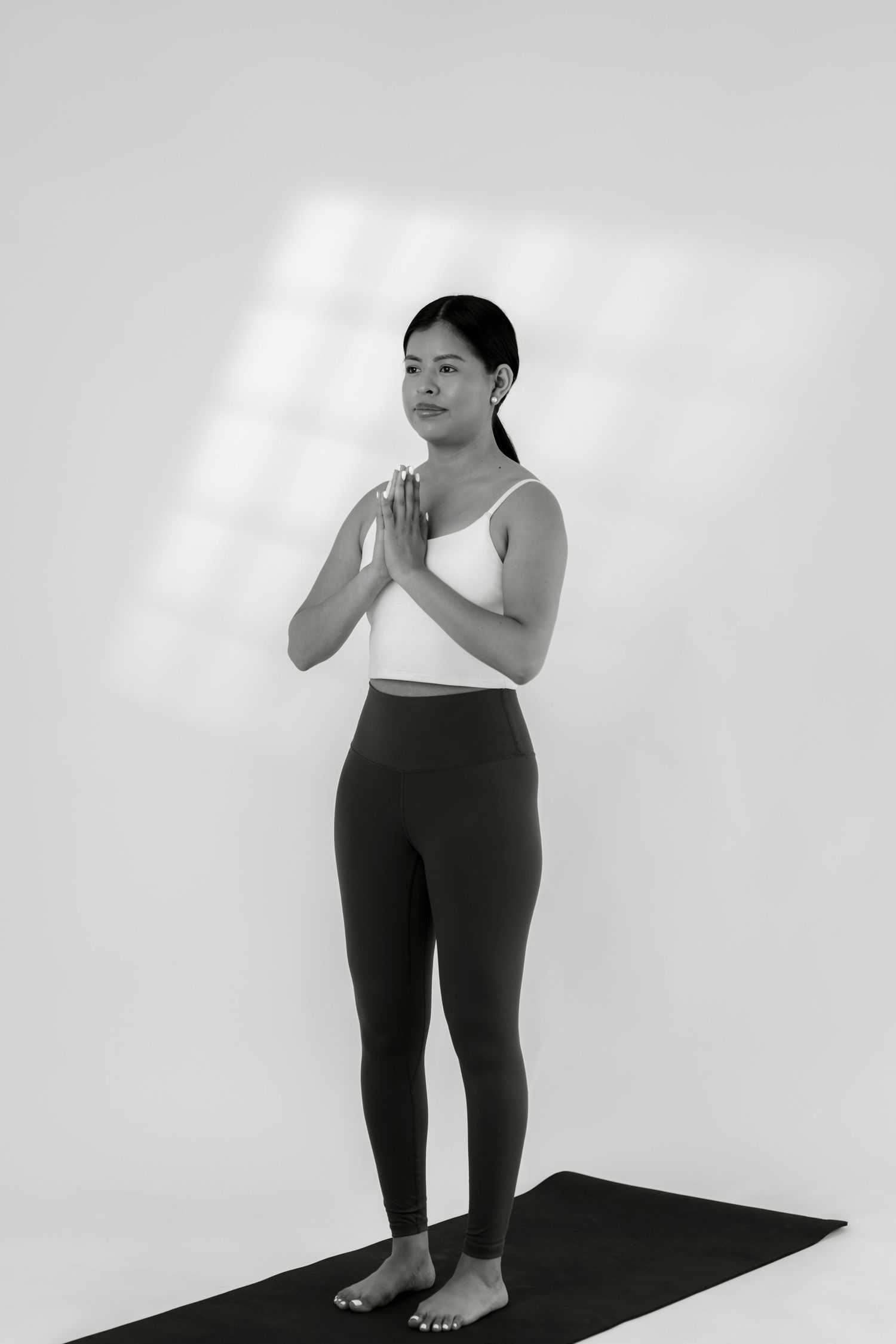 Woman in white sports bra and leggings stands in prayer pose on yoga mat in black and white photo.