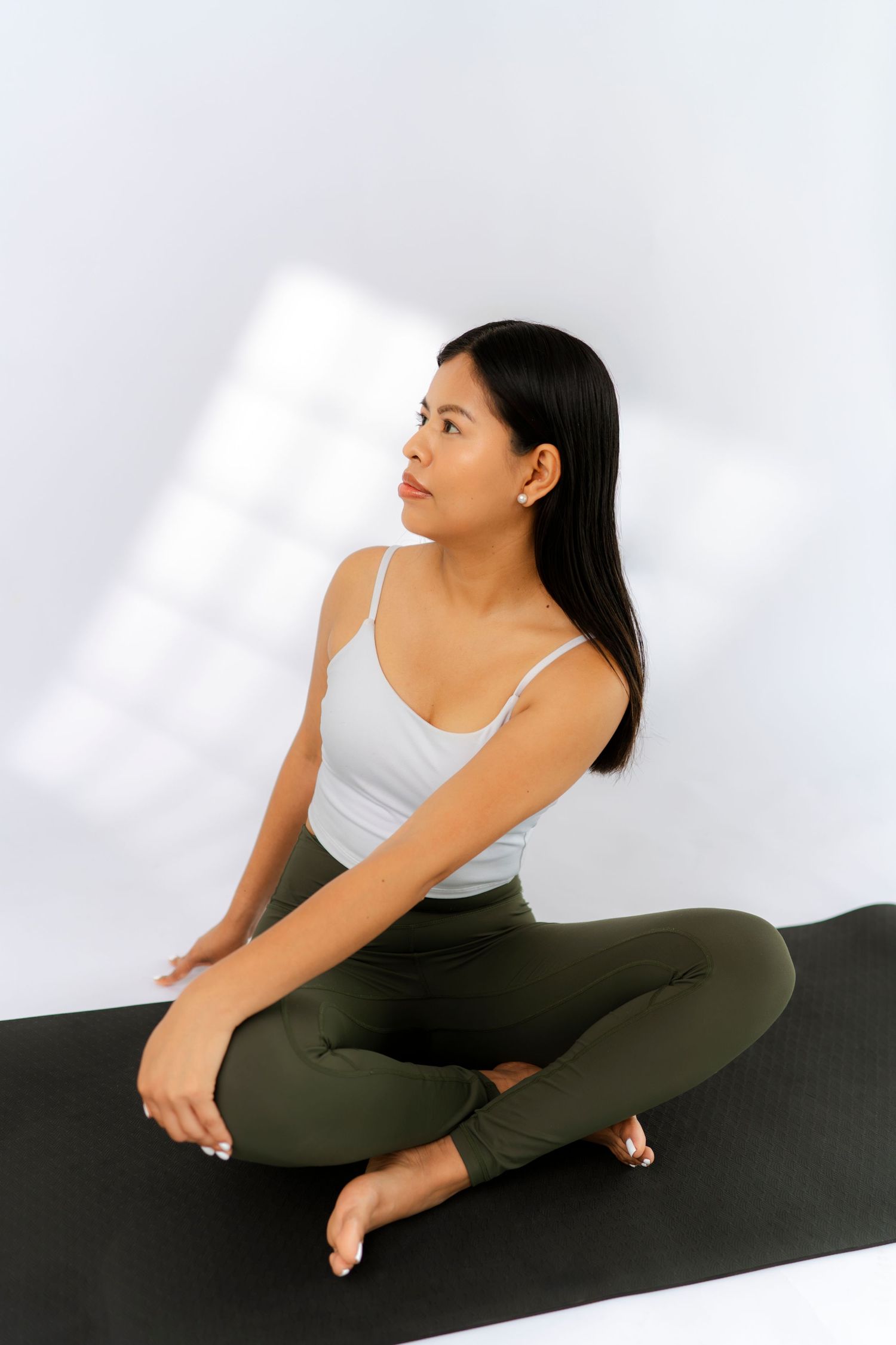 Young woman in white tank top and green leggings sitting cross-legged on black yoga mat against white wall.