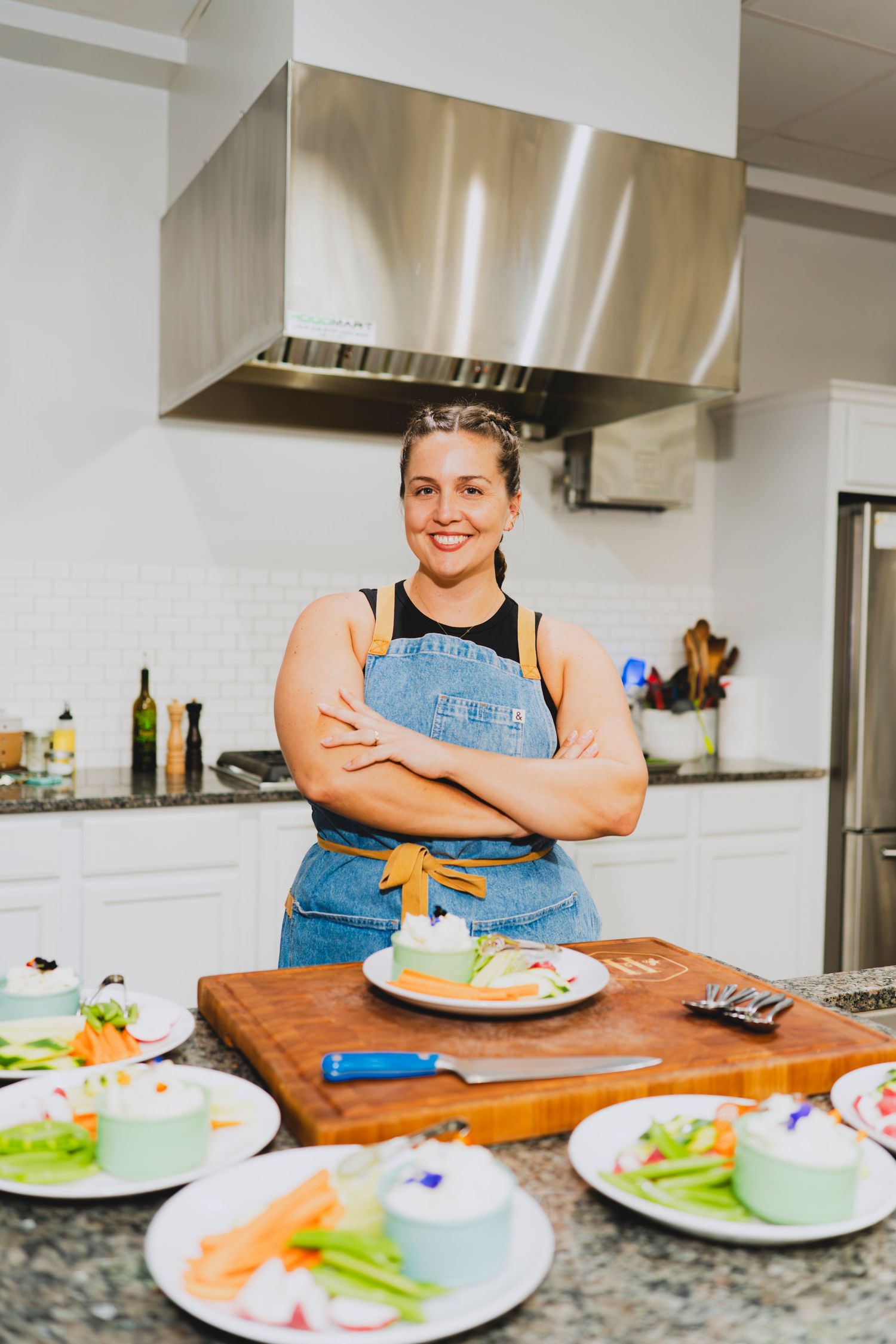Smiling chef in denim apron stands arms crossed behind a wooden cutting board with plated dishes in a modern kitchen.
