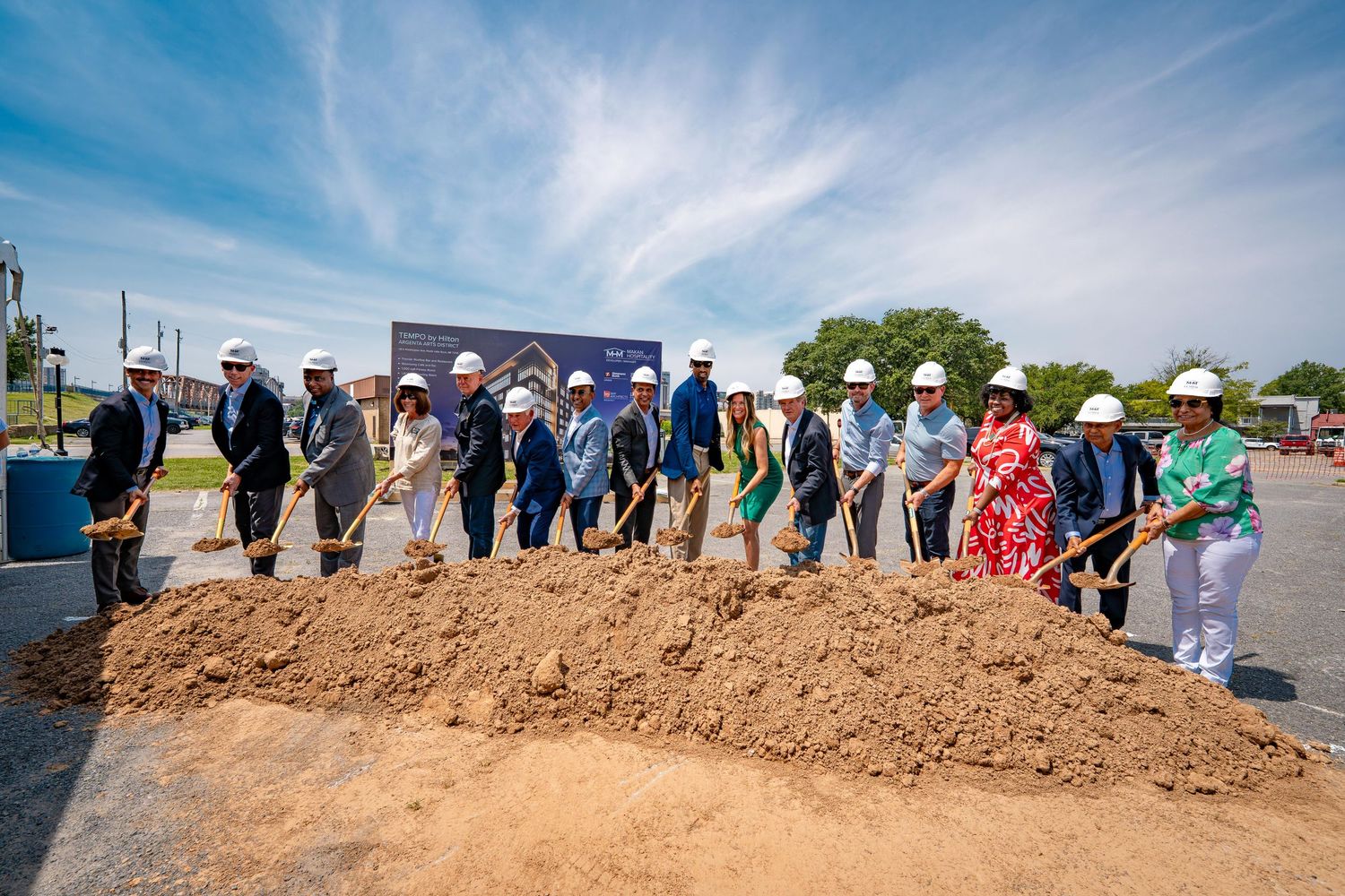 Line of people in hard hats participating in ceremonial groundbreaking with shovels on dirt mound.