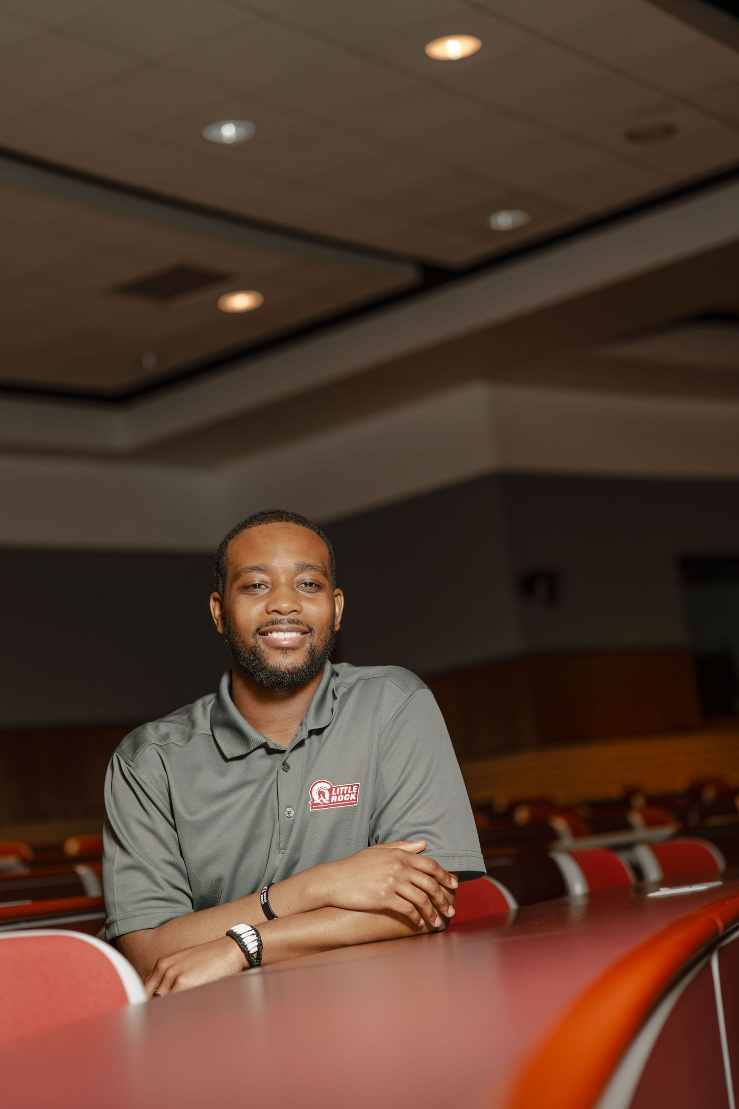 Professional employee in grey polo shirt smiling while seated in a conference room with red chairs.