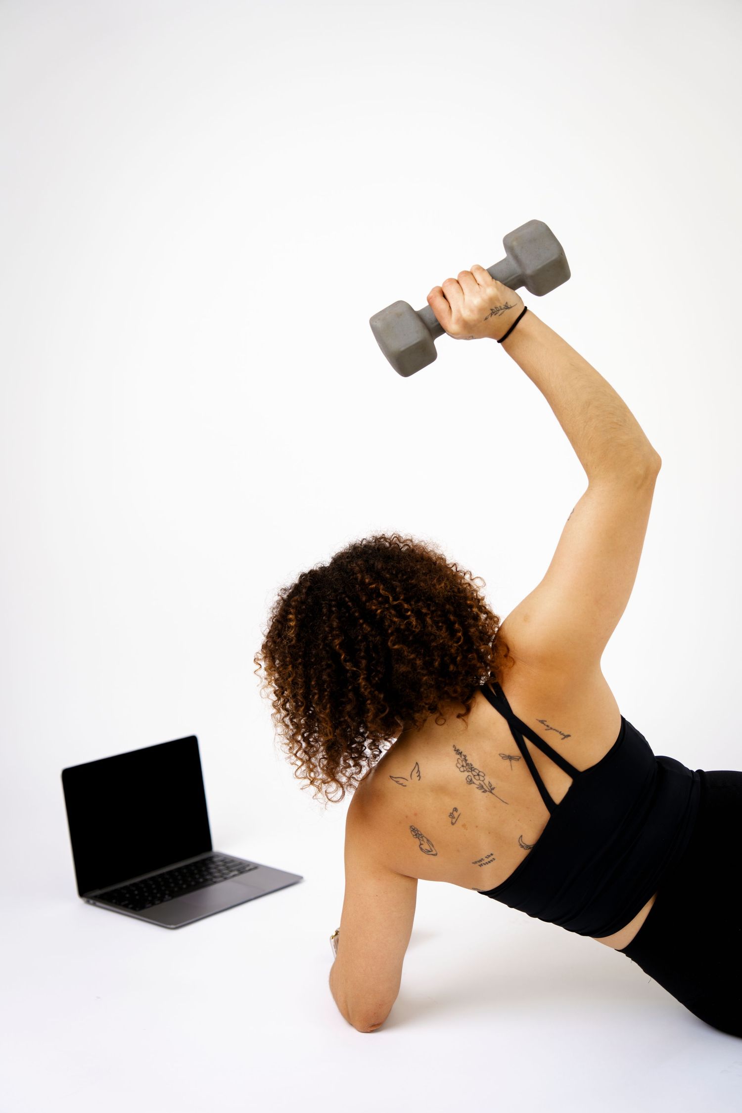Woman with curly hair doing dumbbell exercise while following online workout on laptop, wearing black sports bra.