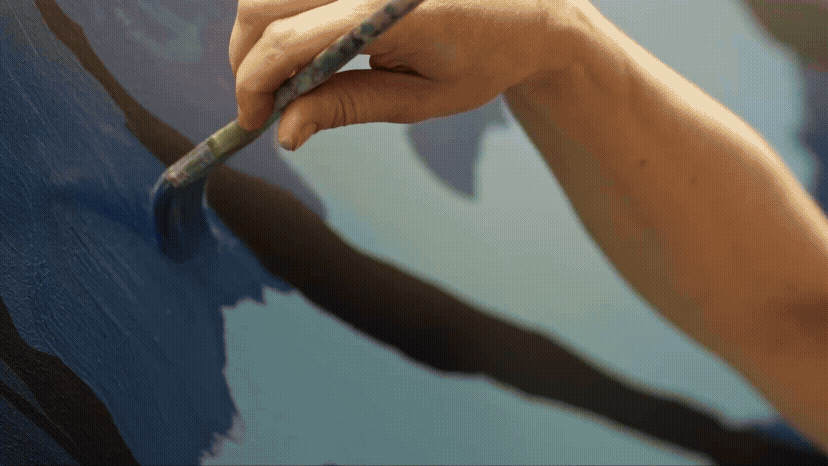 A close-up of an artist's hand painting blue strokes on canvas with a fine brush.