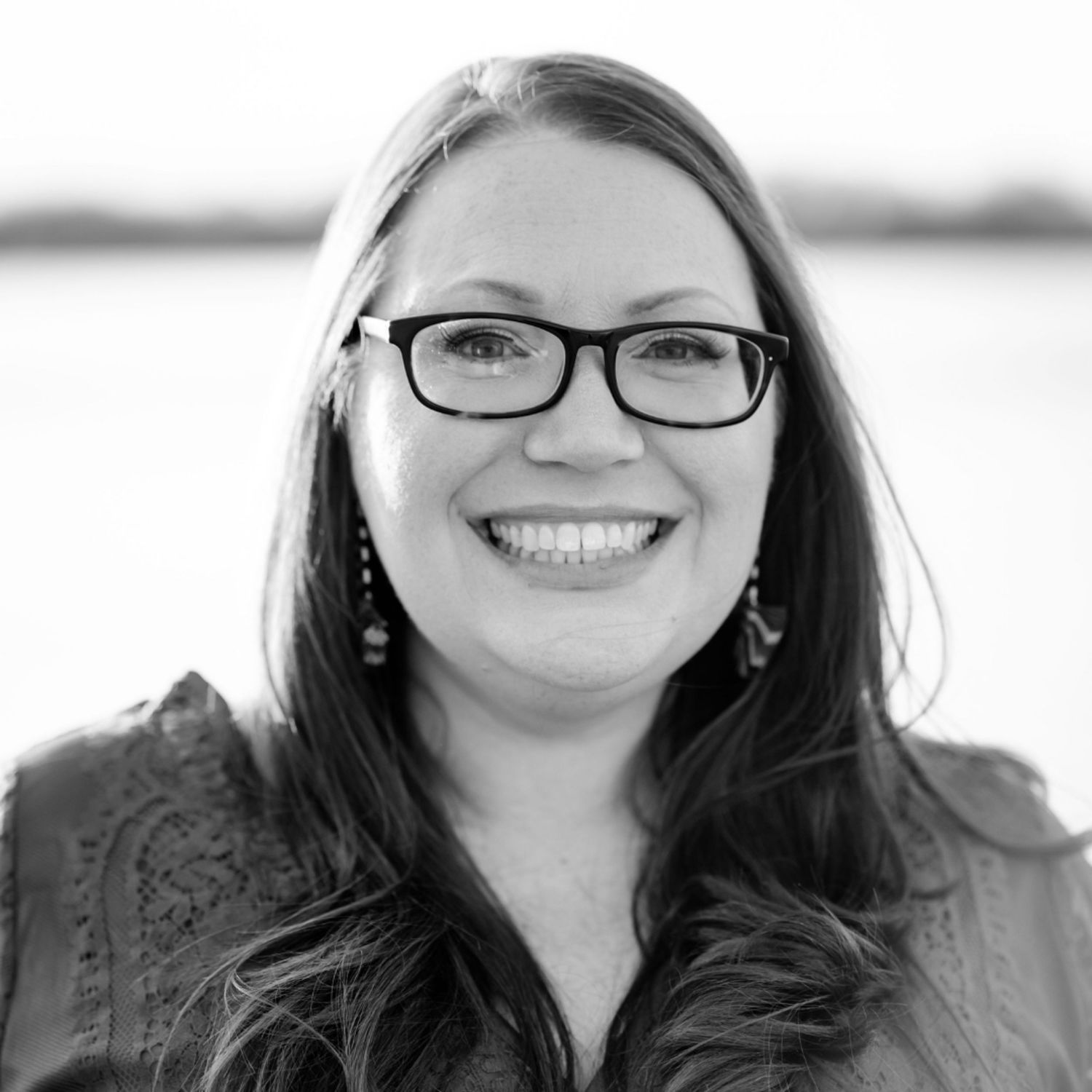 Professional headshot portrait of someone with long dark hair and glasses smiling warmly at the camera.