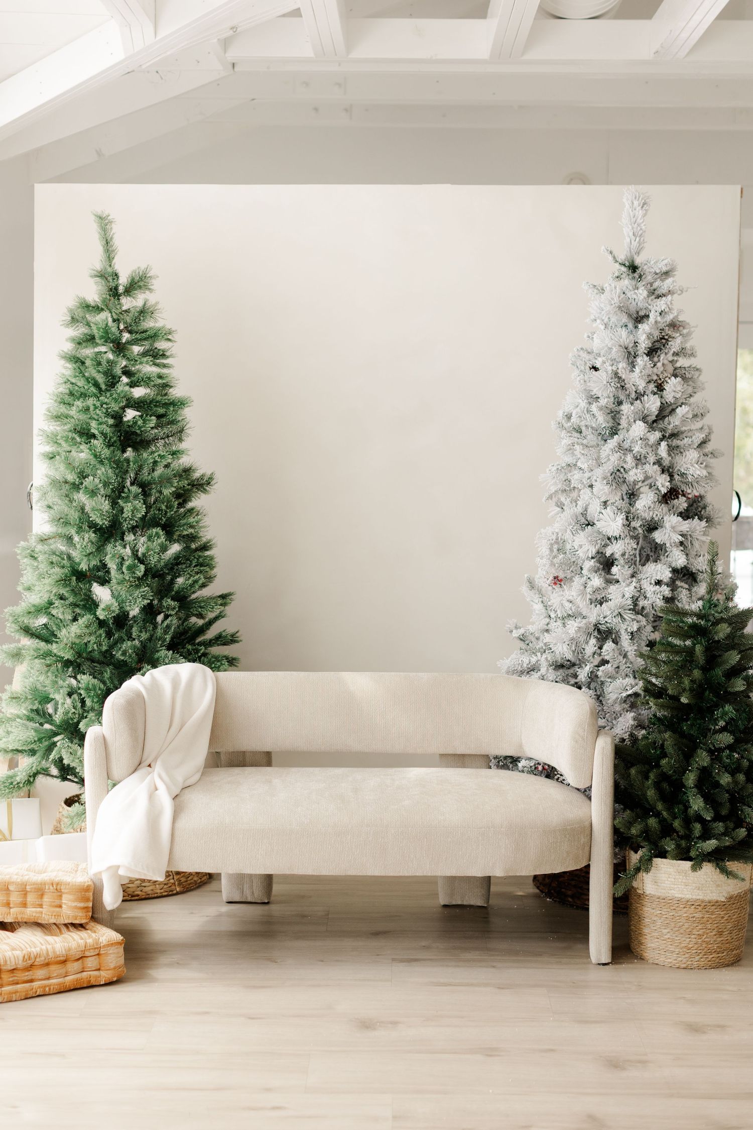 Cozy holiday living room with green and snow-flocked Christmas trees flanking a cream bench with white throw blanket.
