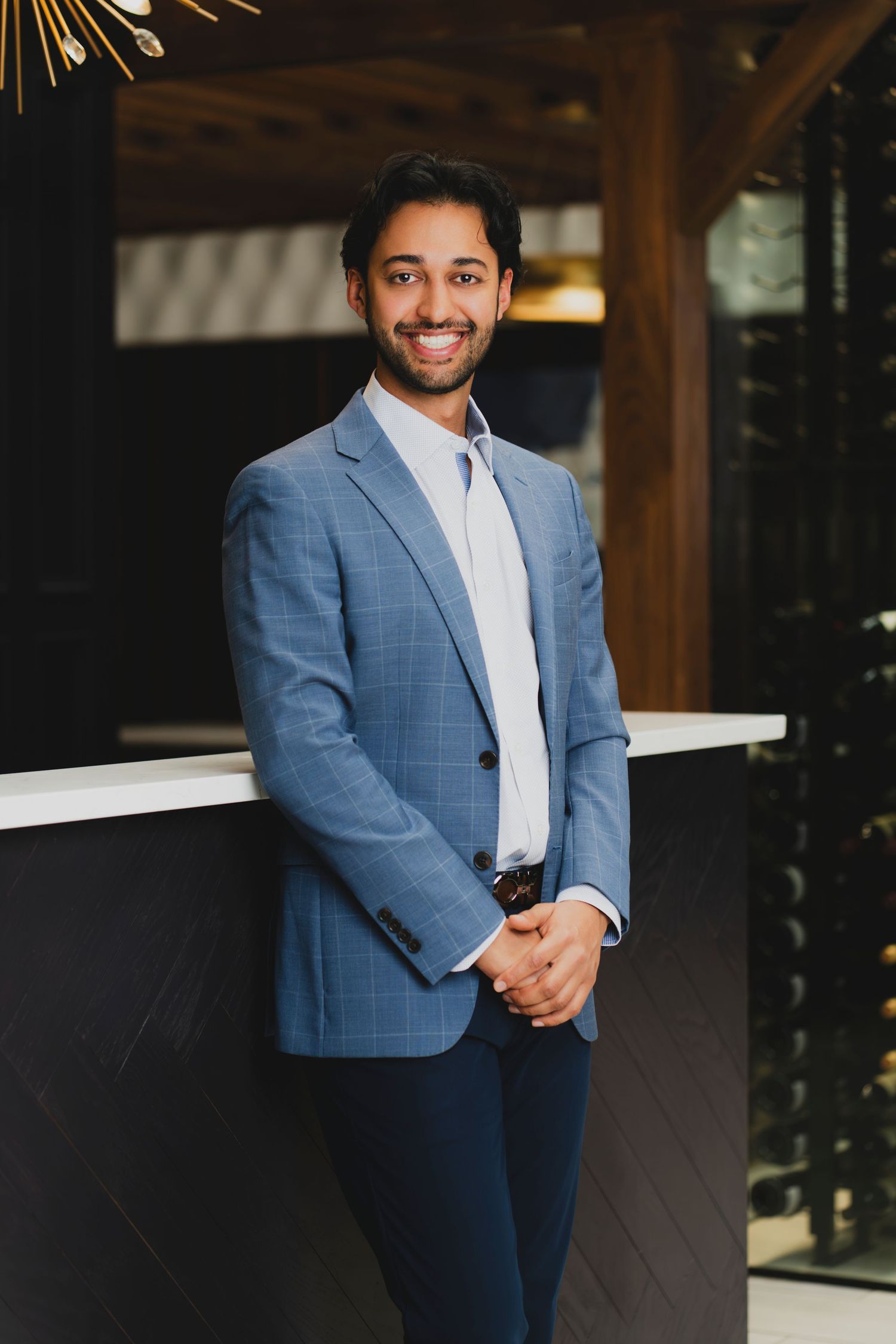 Smiling young professional man in blue plaid blazer posing confidently in modern upscale interior setting.