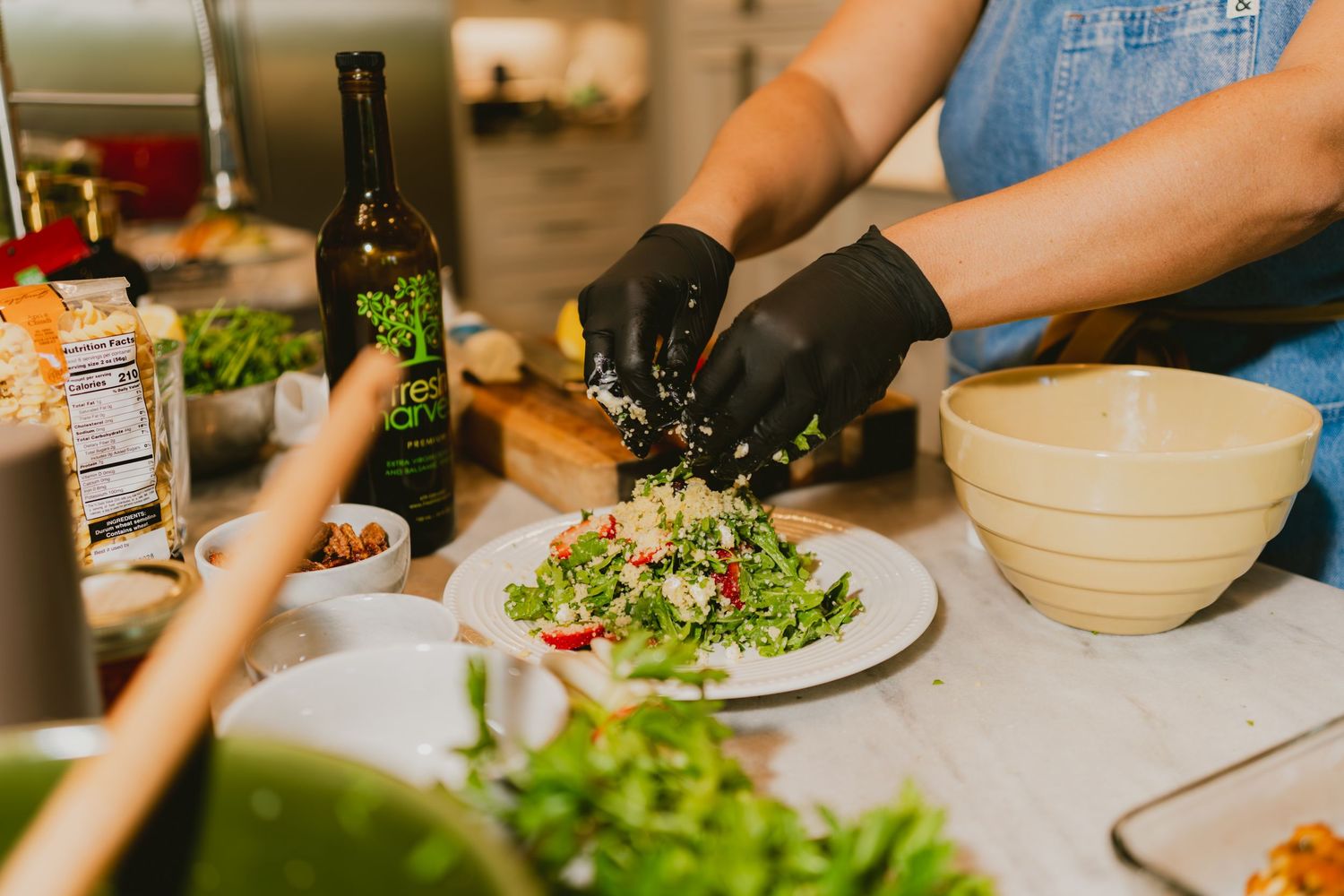 Chef in black gloves plating a fresh green salad on a white dish beside olive oil bottle in kitchen.