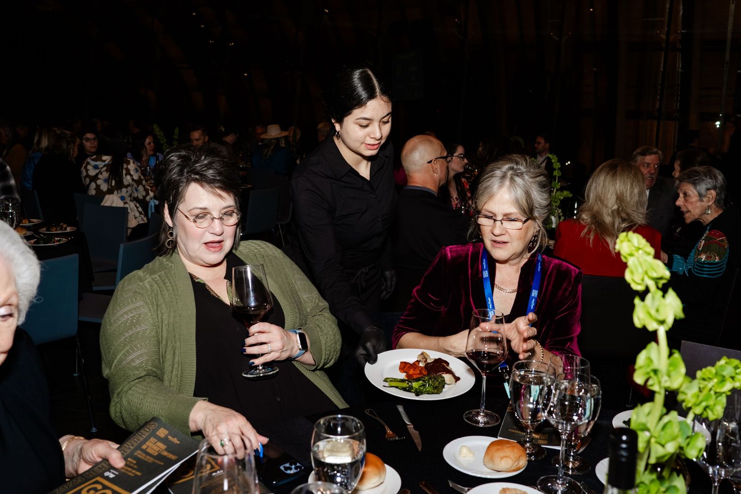 Guests enjoying food and drinks at an elegant evening event with dark lighting and floral centerpieces.