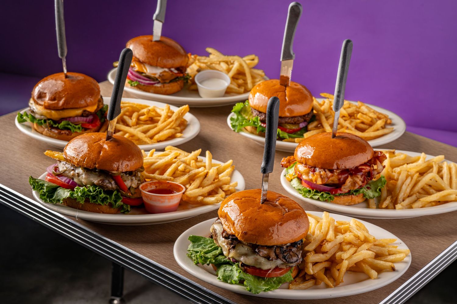 Gourmet burgers with french fries served on dark plates against a purple background.