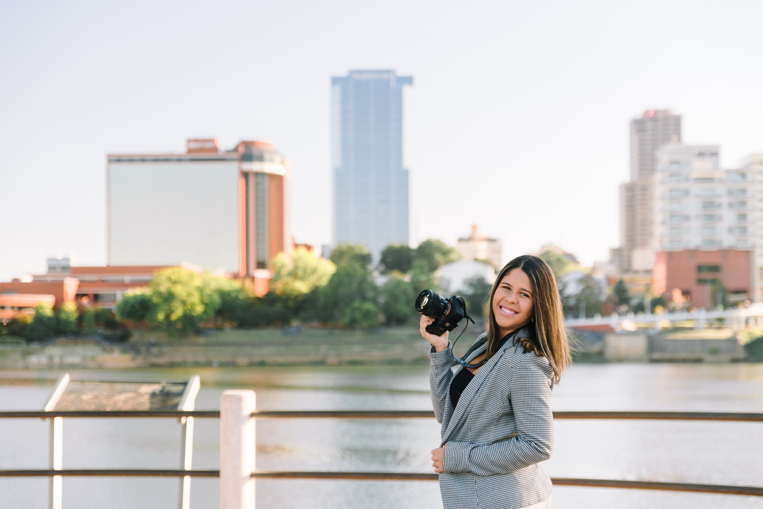 Professional photographer with camera standing on riverfront with city skyline and buildings in background.