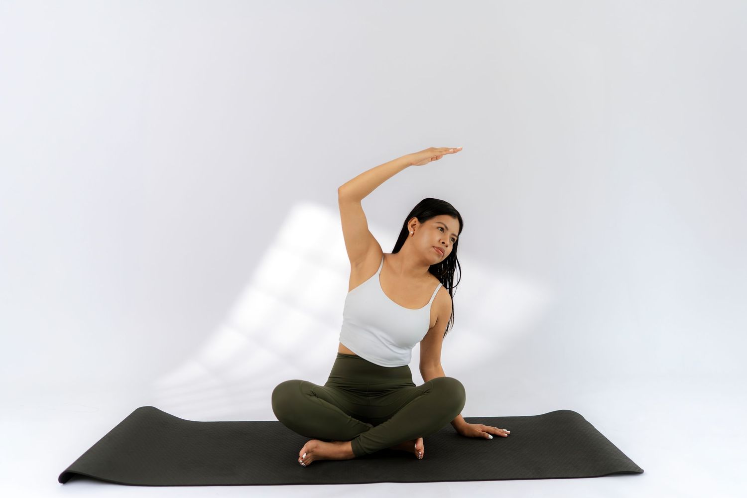 Woman in white top and green leggings practicing yoga side stretch pose on black mat against white background.