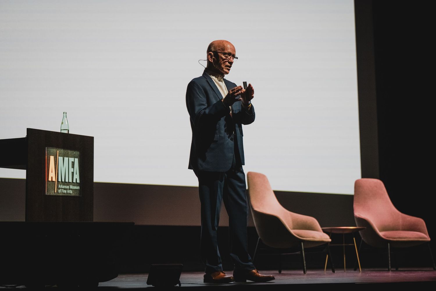 Speaker gesturing while presenting on stage with pink chairs in background.