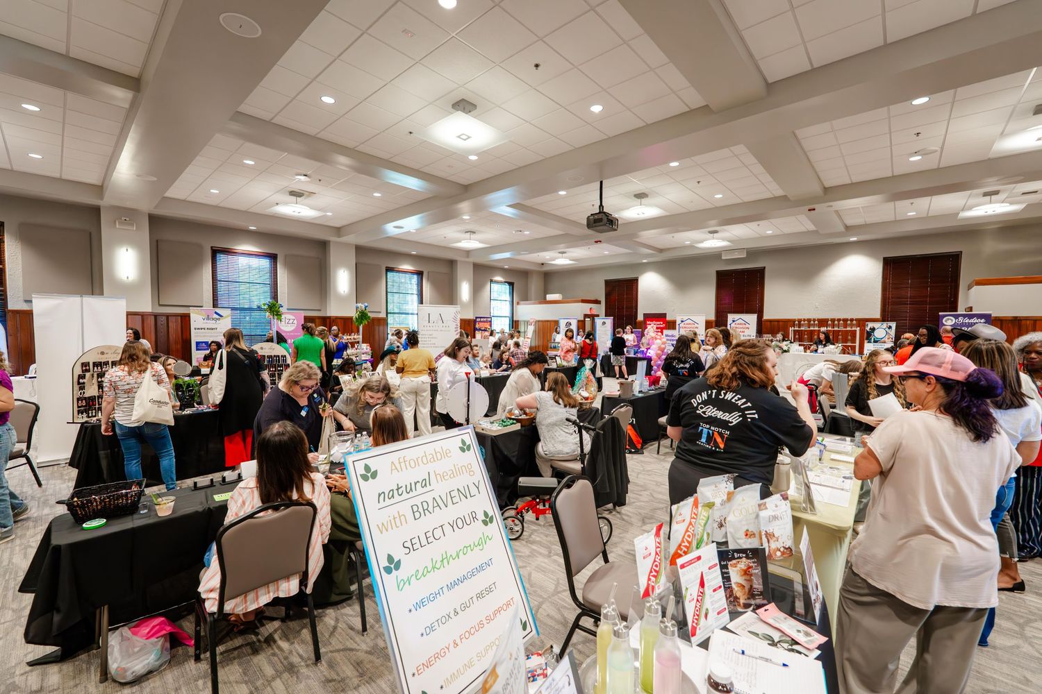 Wide panoramic view of a crowded indoor event space with multiple vendor tables and displays.