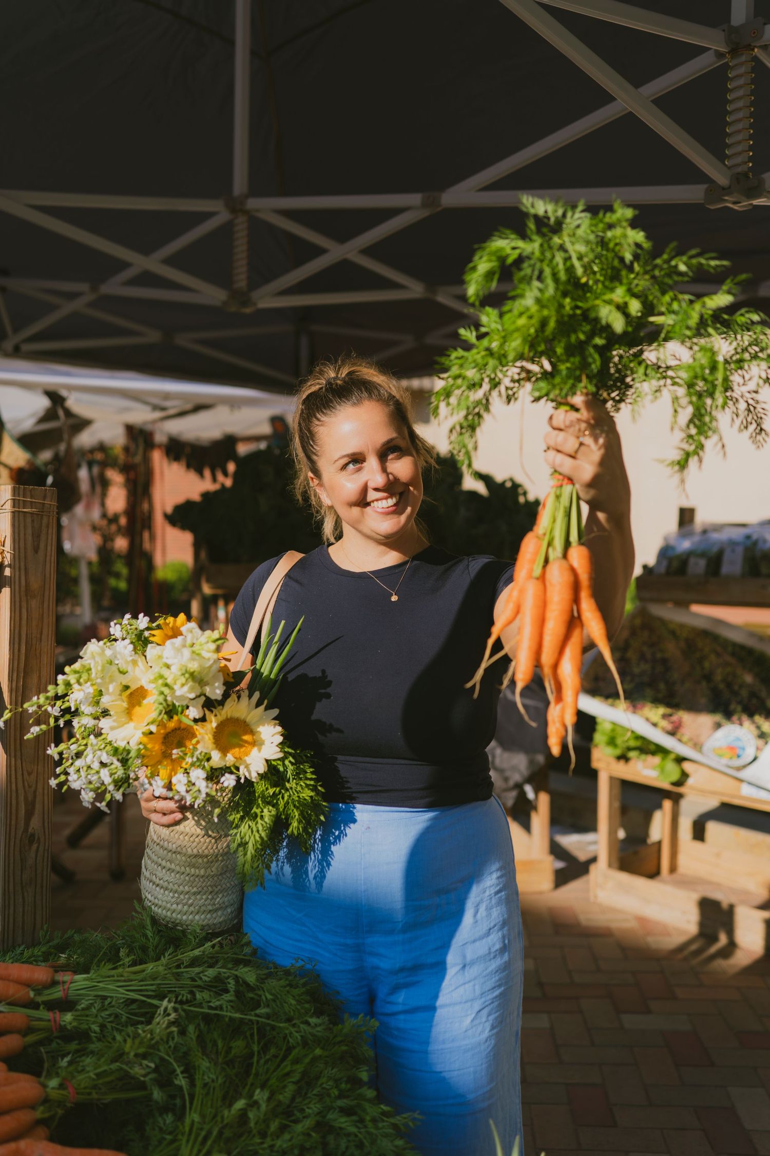 Smiling woman holding fresh carrots and wildflower bouquet at outdoor farmers market in blue outfit.