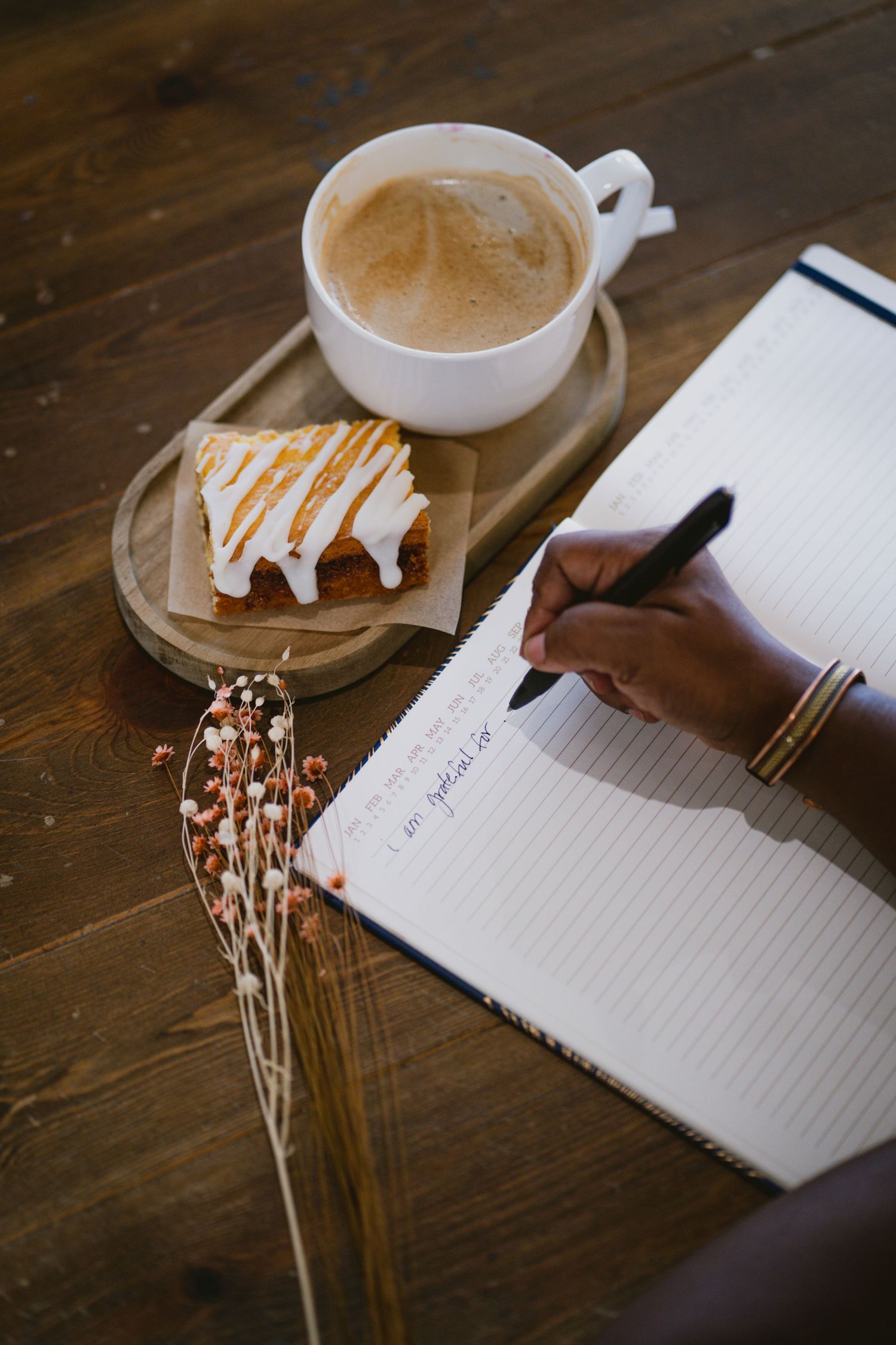 Person writing in notebook with coffee cup and dried flowers on wooden table.