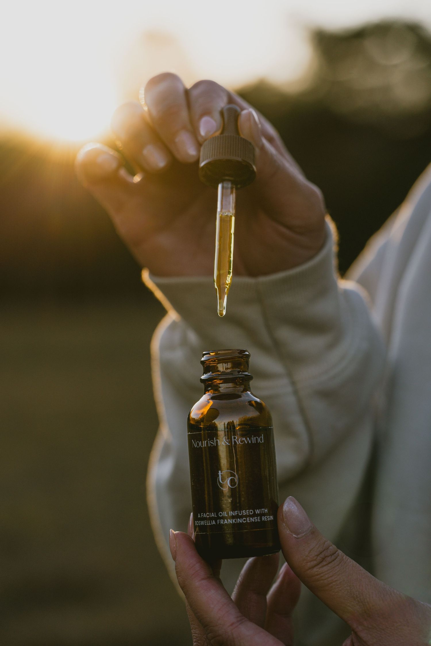 Hands holding glass dropper bottle of skincare serum against sunset background.