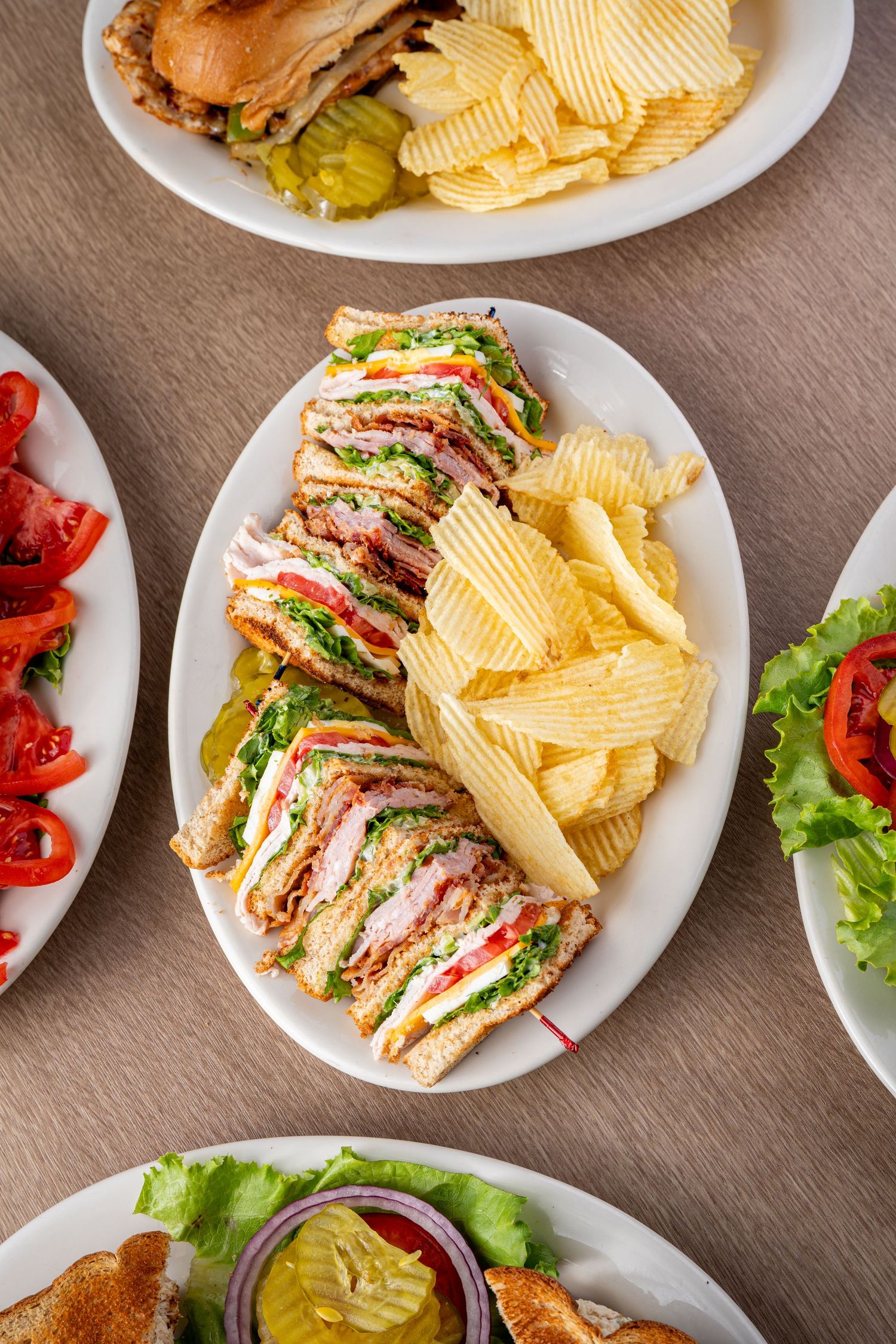Multiple club sandwiches served on white plates with potato chips and fresh vegetables.