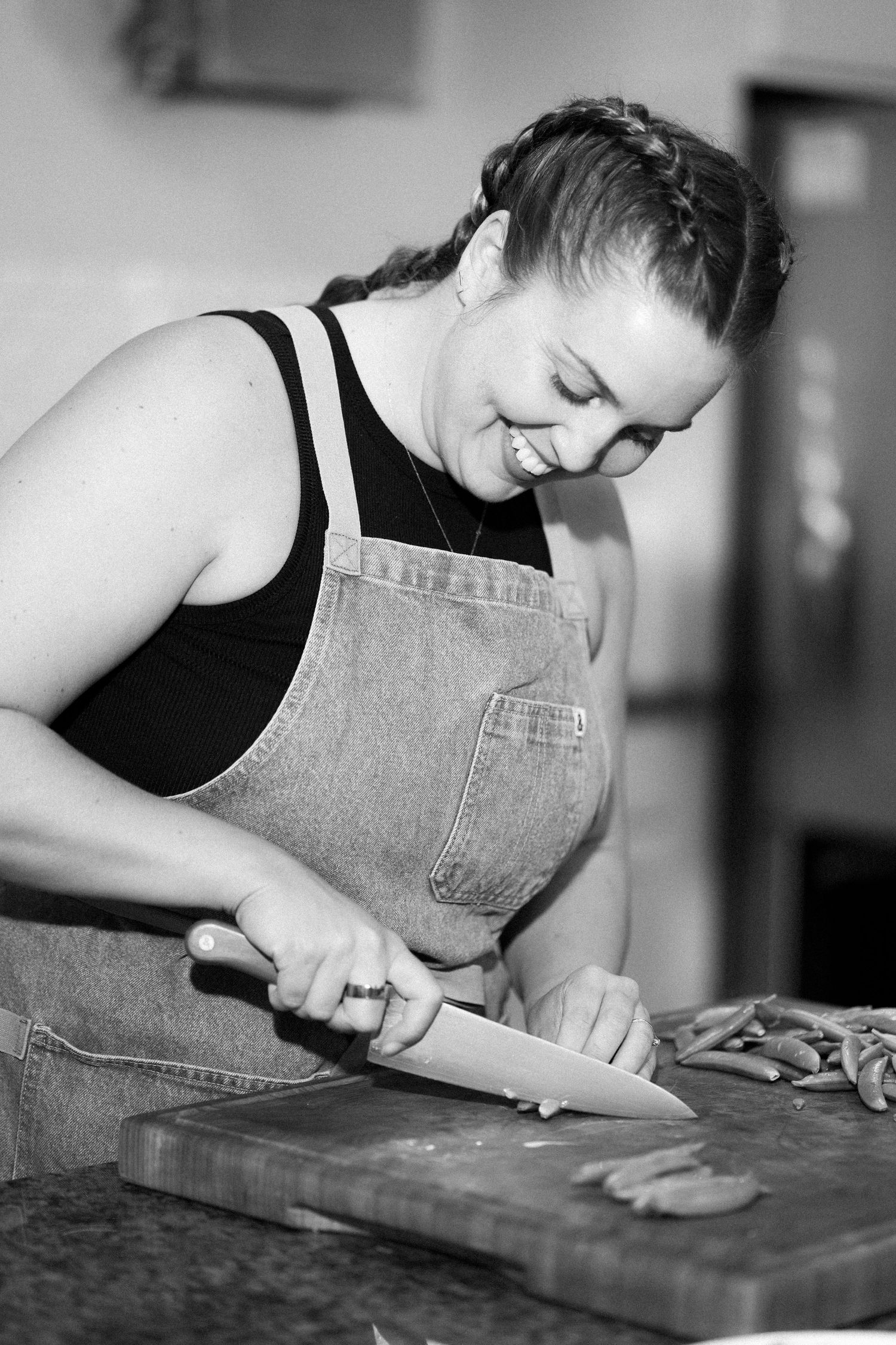 Smiling woman in denim apron chopping vegetables on wooden cutting board in black and white kitchen photo.