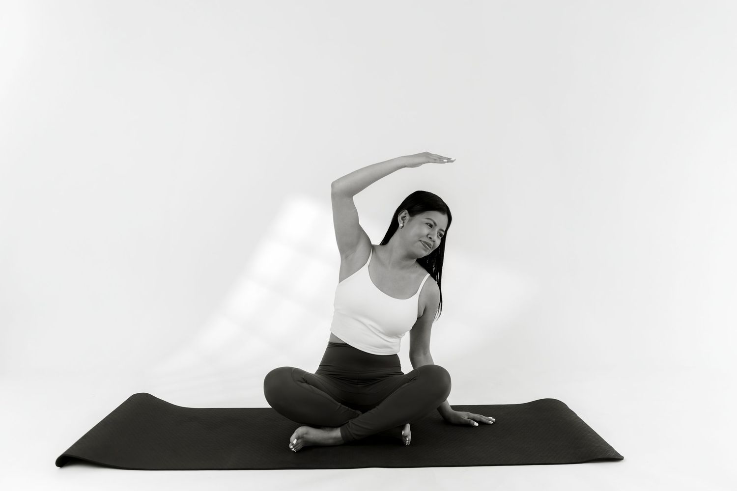 Woman practicing yoga in a seated side stretch pose on a black mat, black and white studio photo.