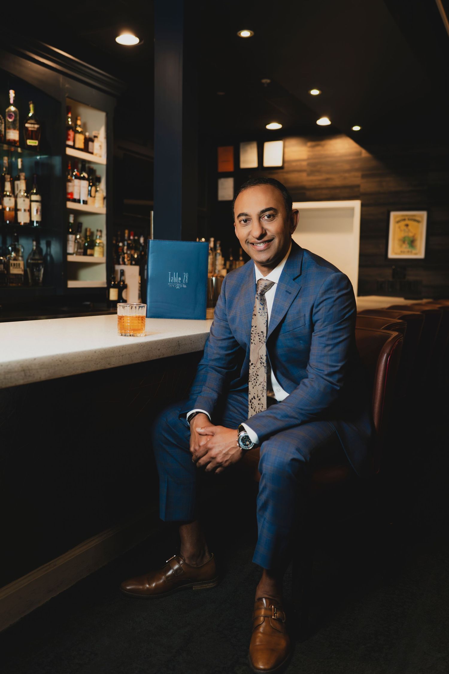 Man in blue suit sitting at upscale bar with whiskey glass and menu, smiling confidently.