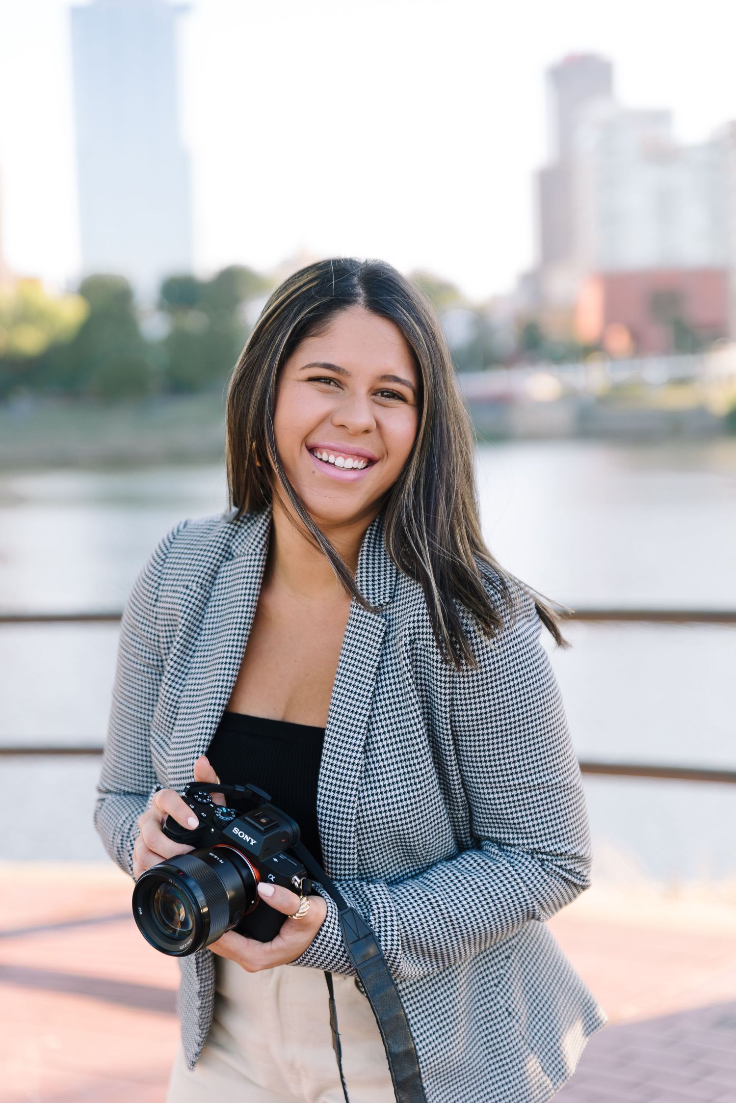 A photographer holding a professional camera against an urban waterfront backdrop.