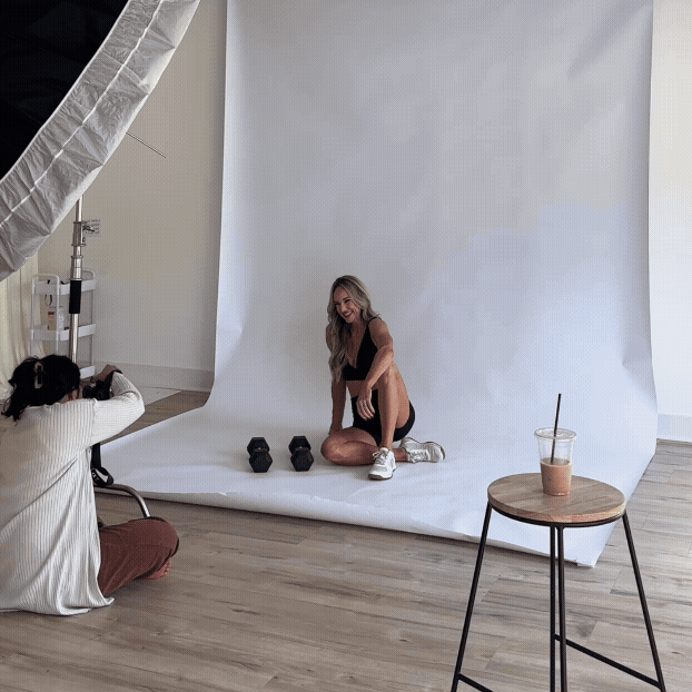 A model poses on a white backdrop during a professional photo shoot while a photographer captures the session in a bright studio.