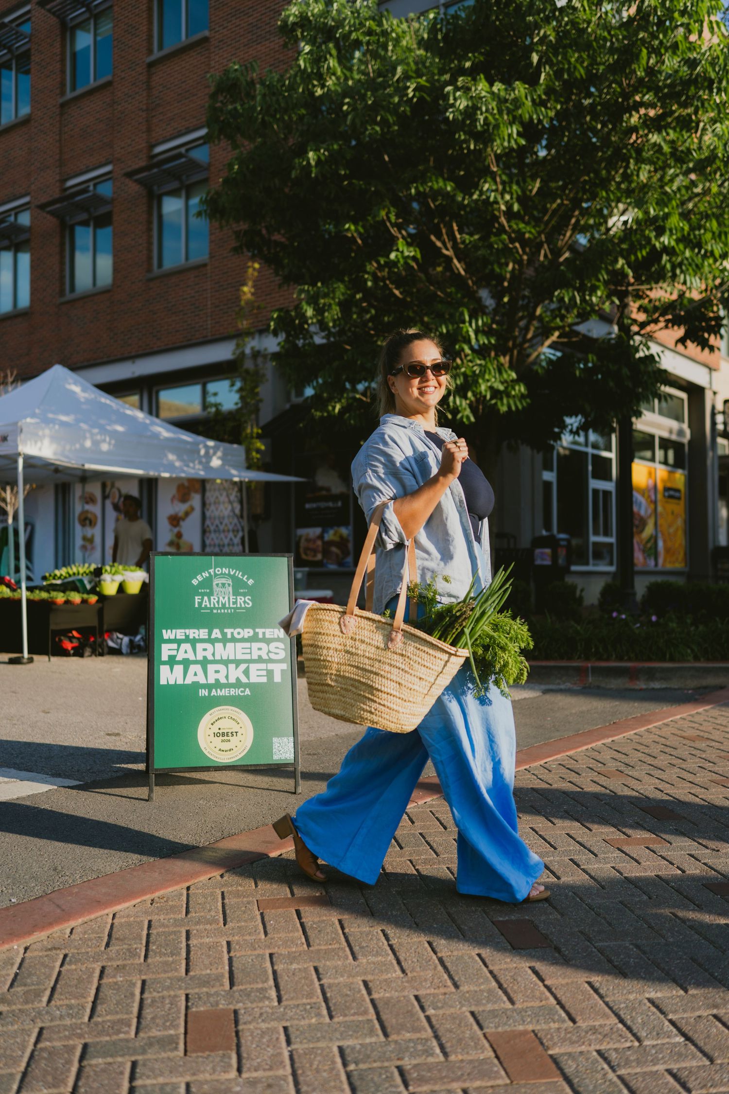 Woman in blue wide-leg pants carrying a straw tote bag at a top-ten farmers market on a sunny day.