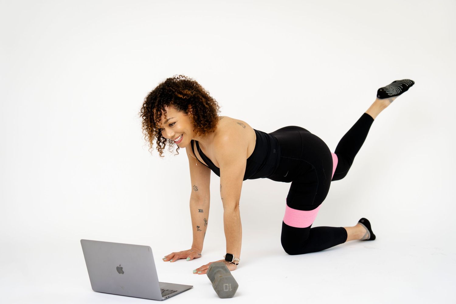 Woman in black and pink workout attire doing donkey kick exercise while watching fitness tutorial on laptop.