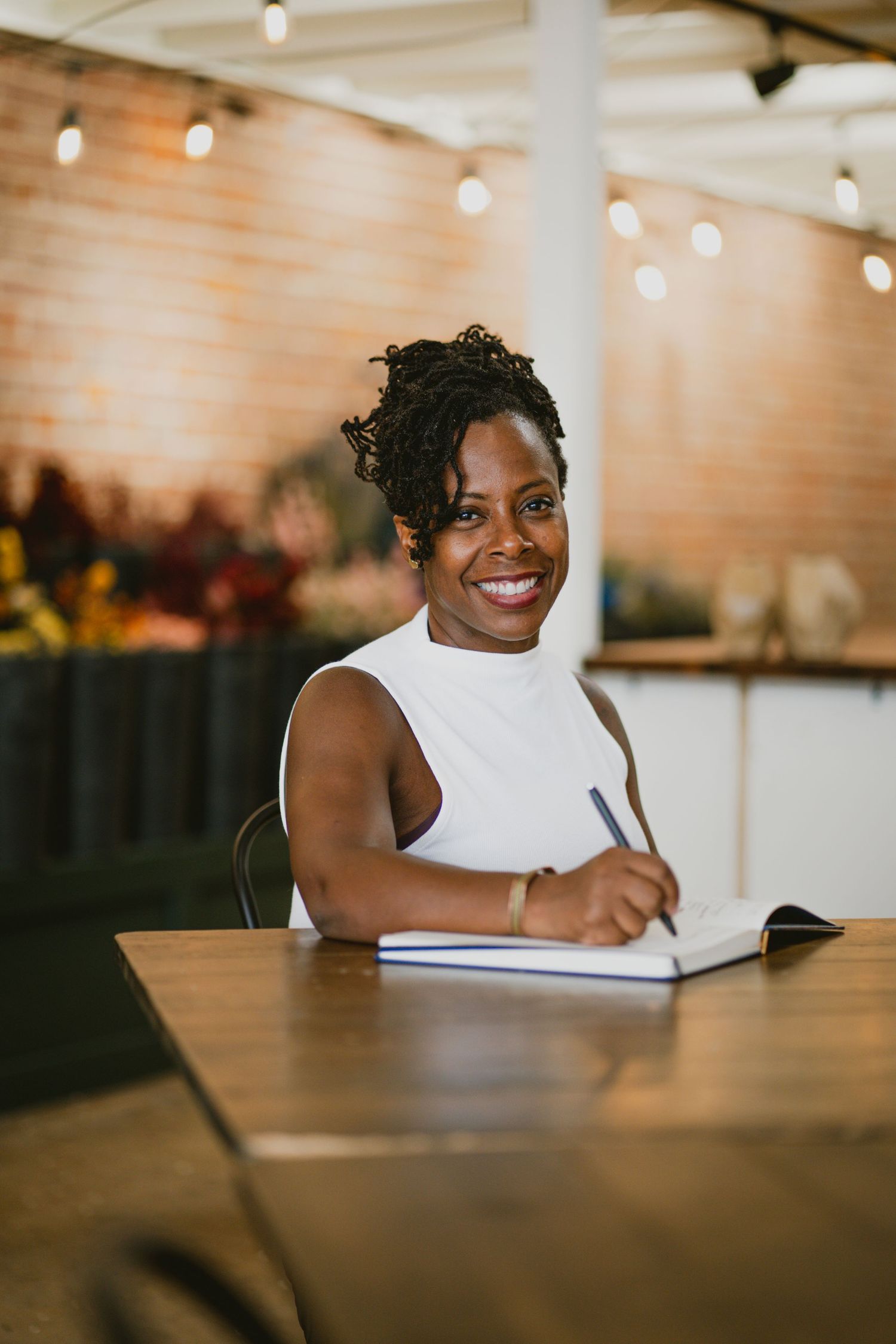 A student writing in a notebook at a wooden table with string lights in the background.