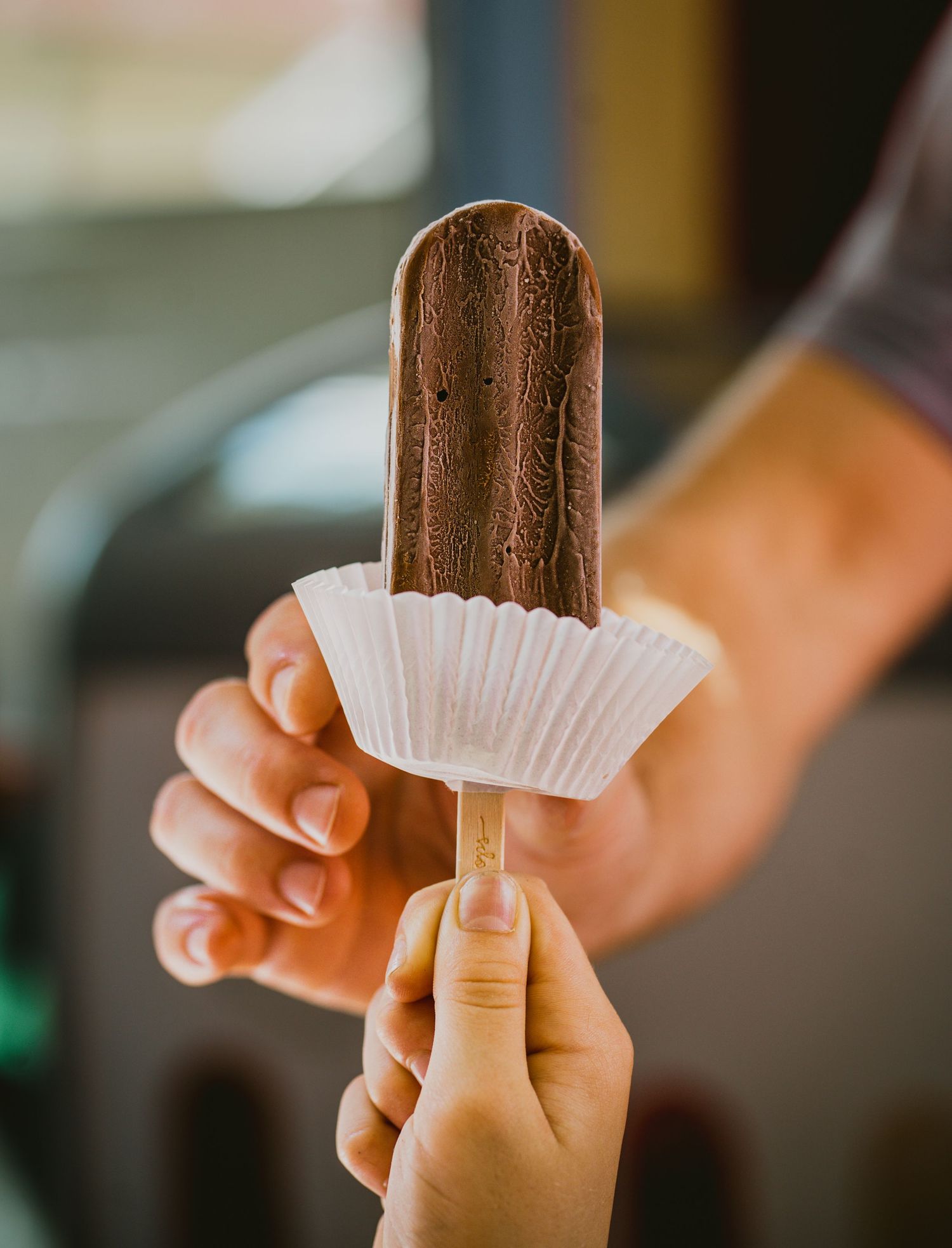 Hand holding a chocolate ice cream bar with paper wrapper.