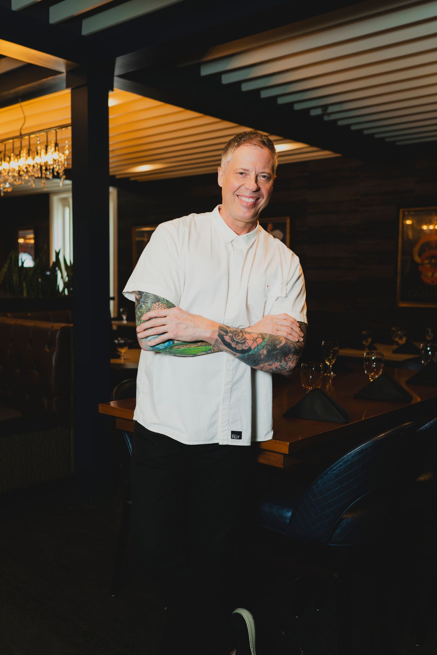 Smiling chef with tattoos in white uniform stands arms crossed in a stylish restaurant interior.