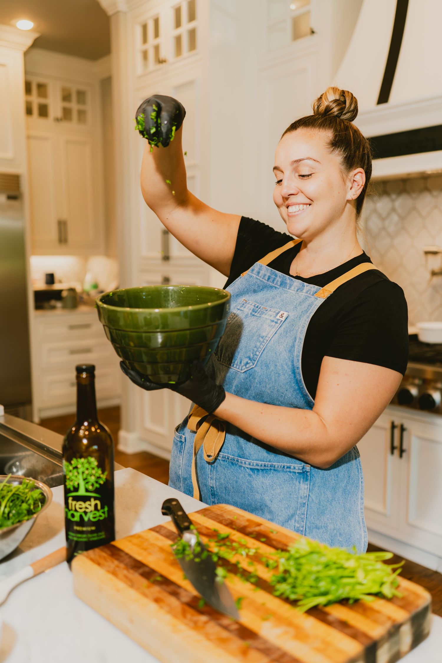 Woman in denim apron tossing fresh greens in green bowl at kitchen island with Fresh Harvest bottle.