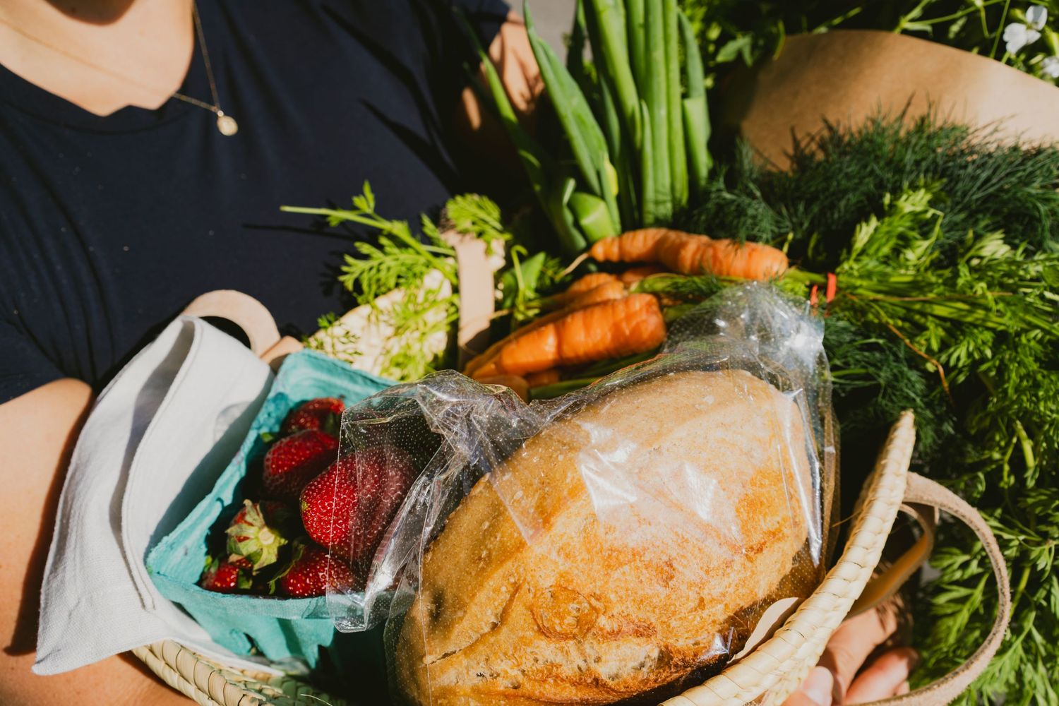 Person holding a wicker basket filled with fresh groceries including strawberries, carrots, bread, and greens outdoors.