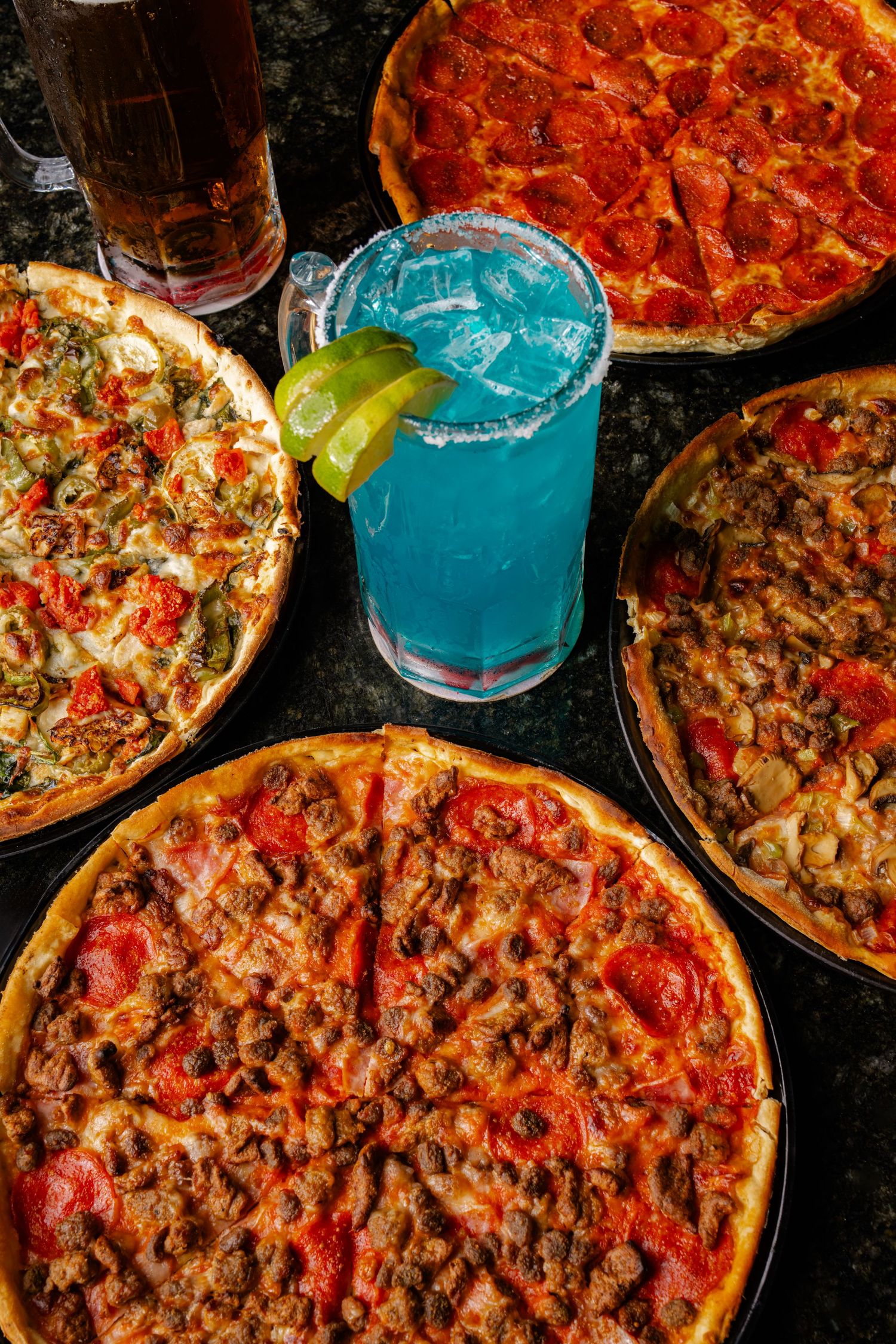 Four pizzas and blue cocktail on dark table with pepperoni and various toppings.
