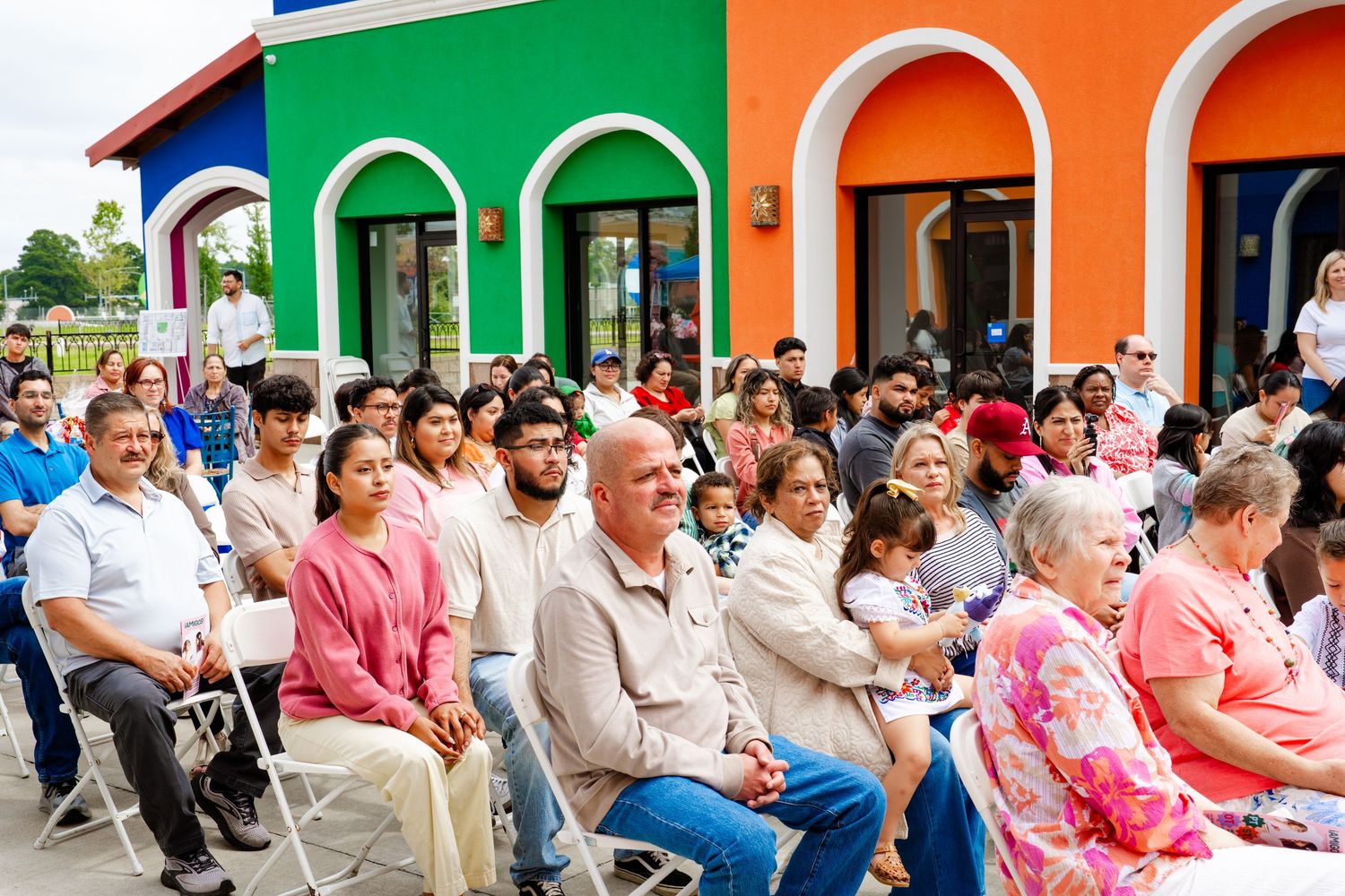 Crowd seated at an outdoor event with orange and green building facade visible in background.