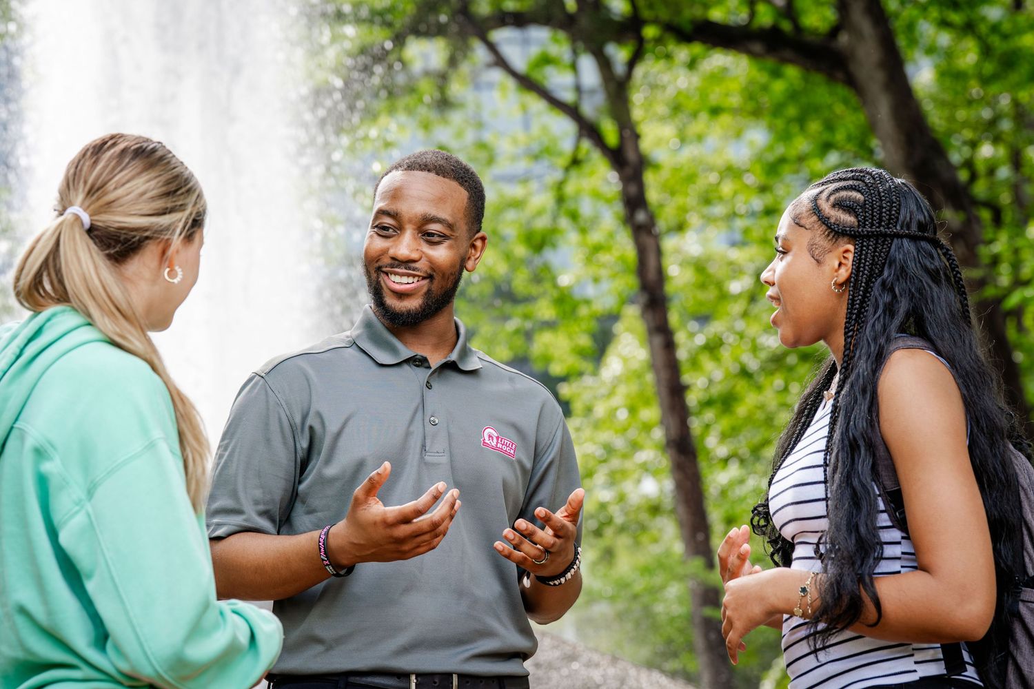 Group of people interacting outdoors near a fountain with trees in background.