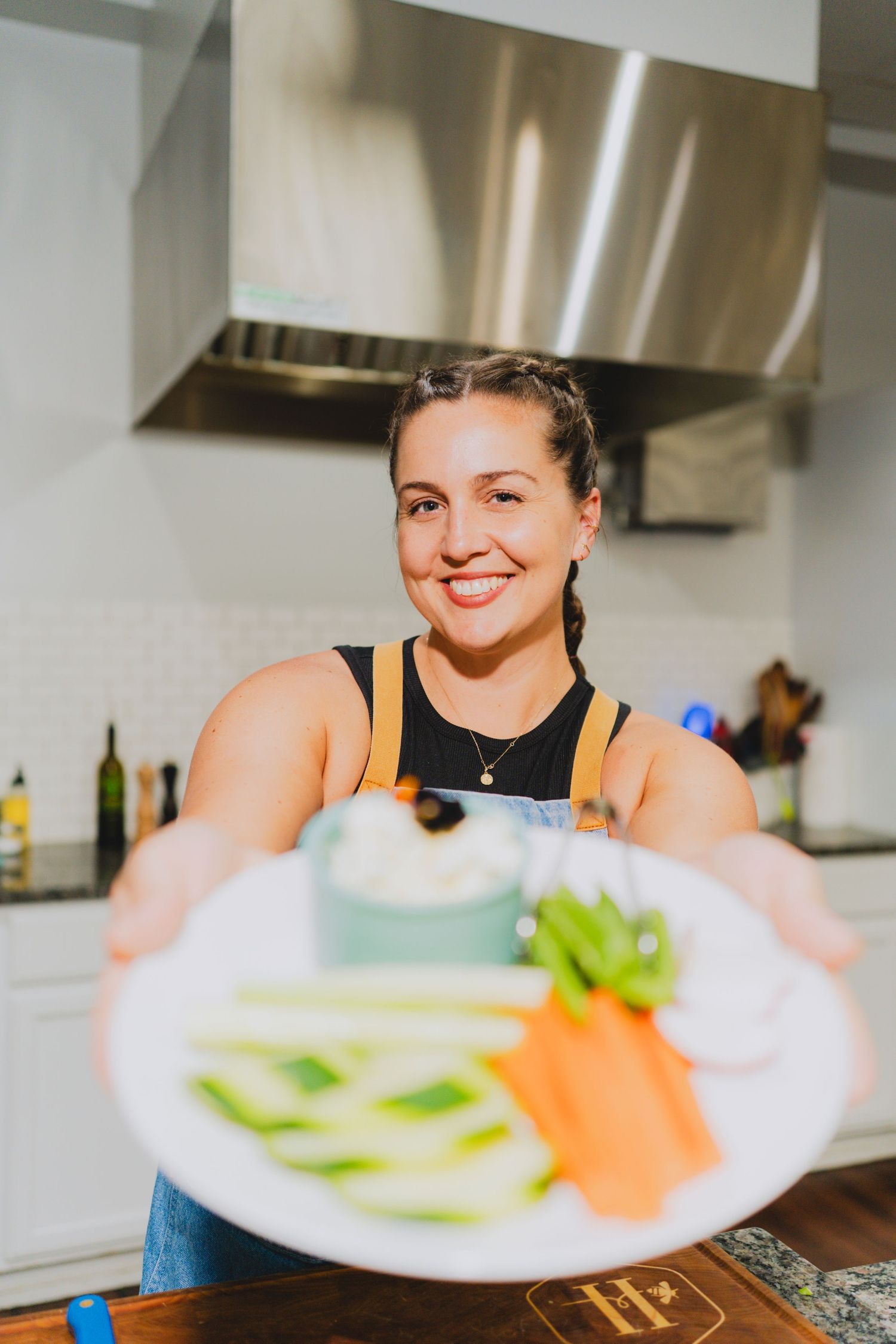 Smiling woman in kitchen holding a white plate with fresh vegetable slices and dip toward the camera.